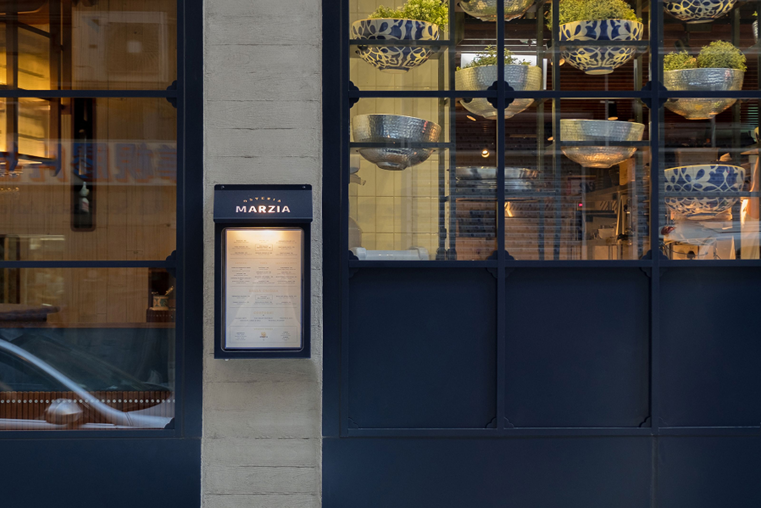 Osteria Marzia exterior with a lit menu board, large windows, and interior decorative bowls.