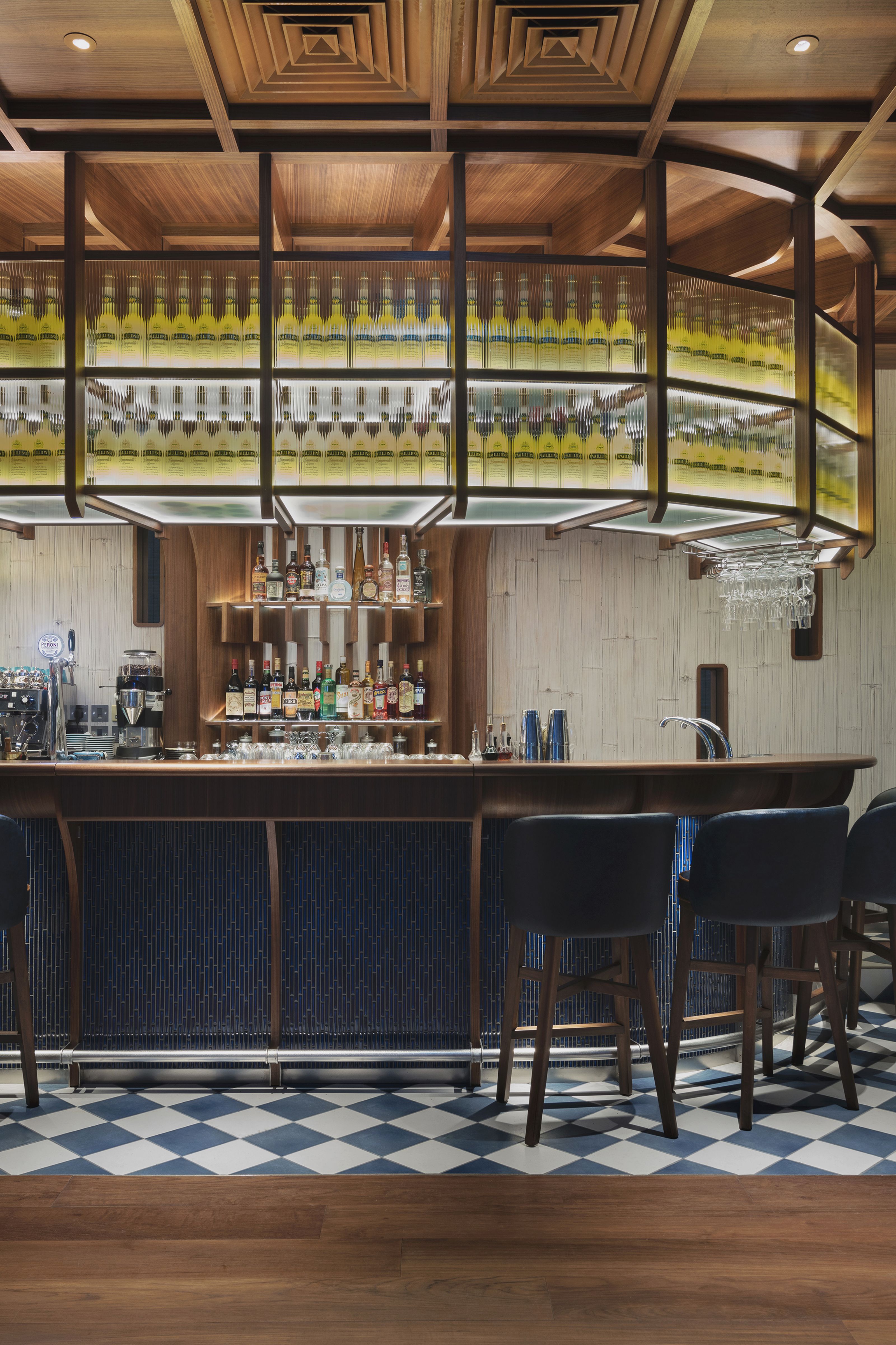 A modern bar interior with a wooden coffered ceiling, a dark wood bar counter with blue paneling, backlit shelves displaying numerous bottles, and a blue and white checkered floor.
