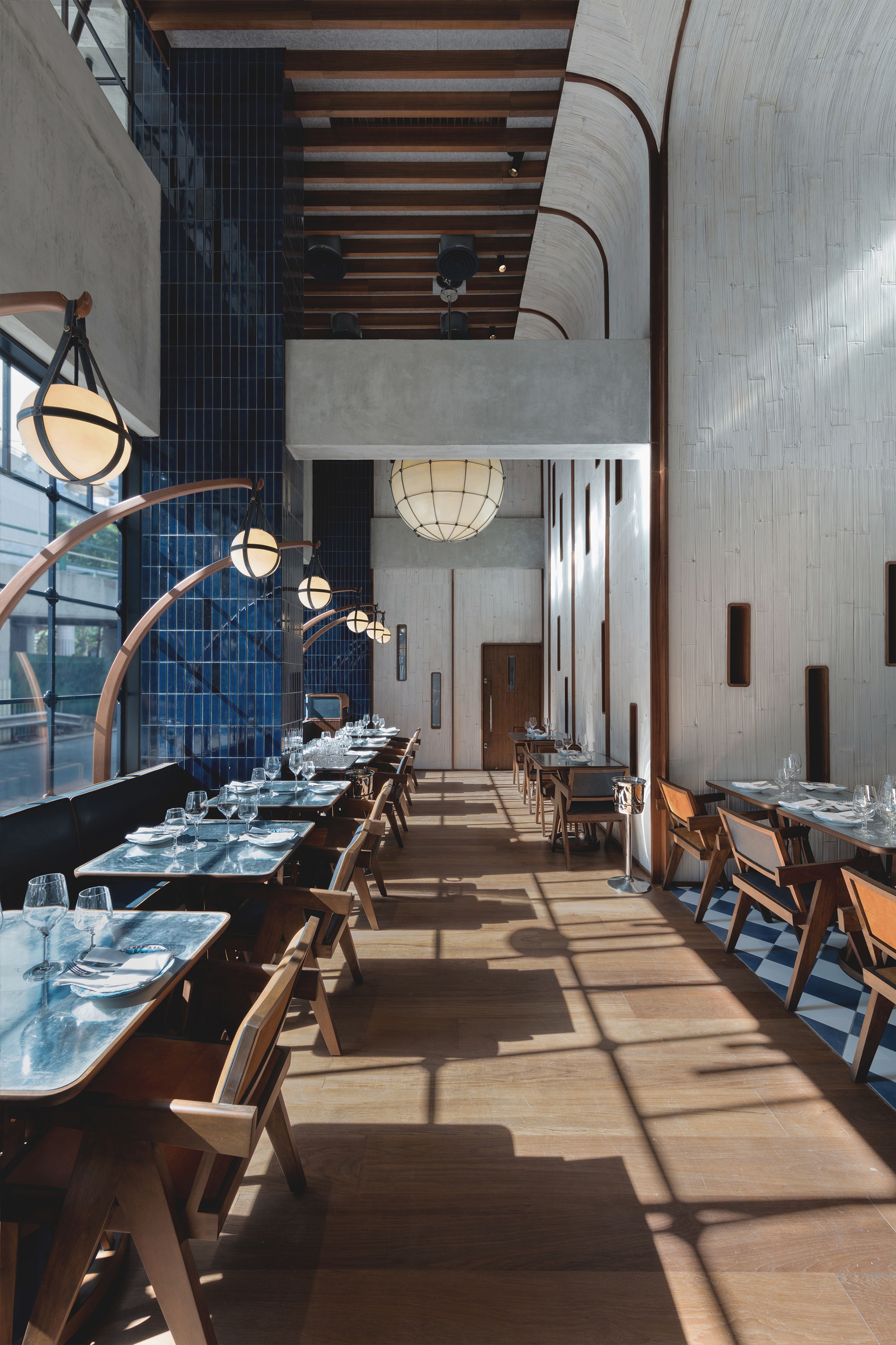 Long modern restaurant dining room with set tables, globe pendant lights, blue tiled walls, and sunlit wooden floors.