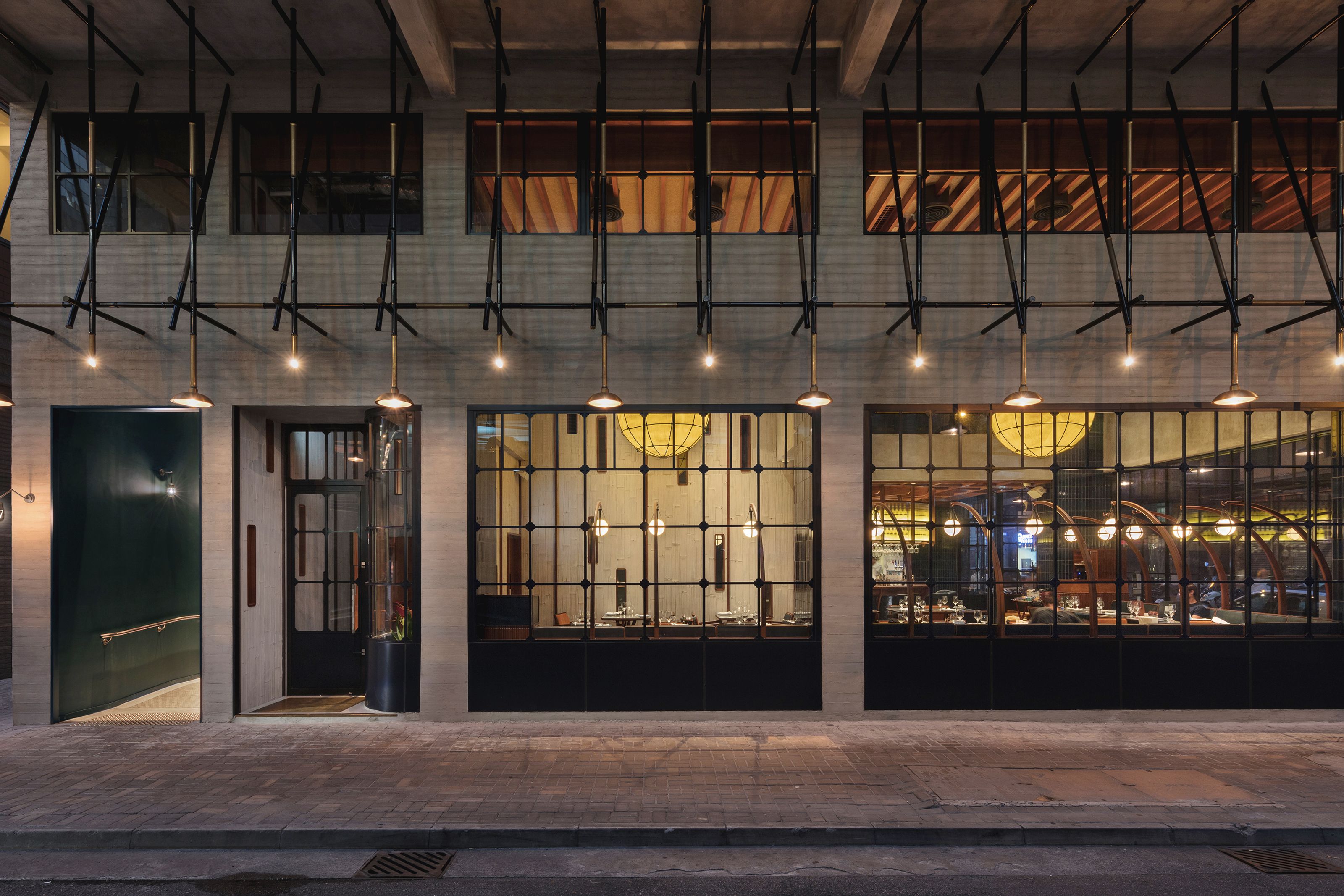 Modern restaurant exterior at night with large grid windows, illuminated interior, and decorative hanging lights.
