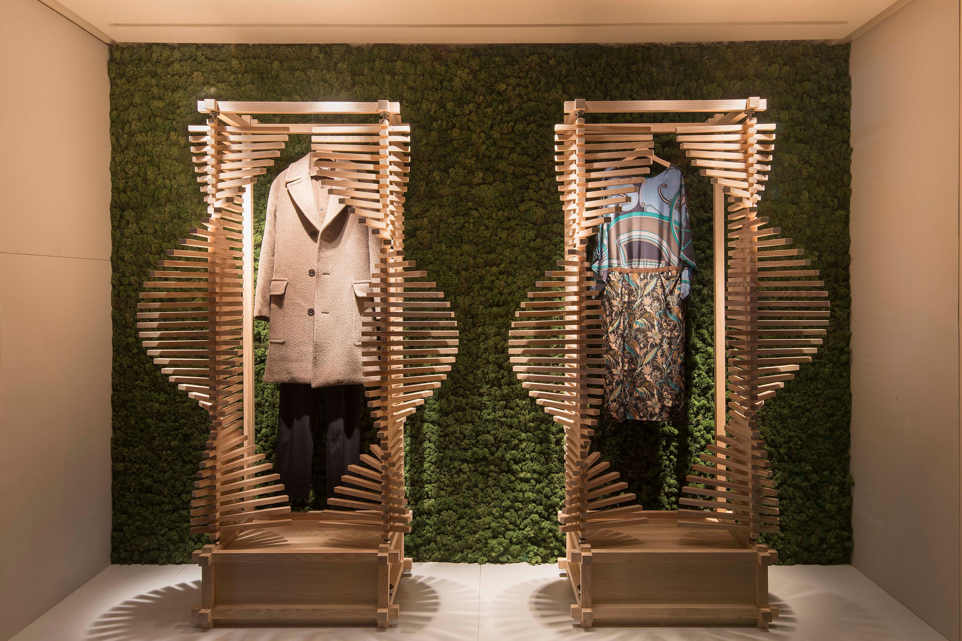 Two spiral-slatted wooden racks display a coat and a dress against a green moss wall.