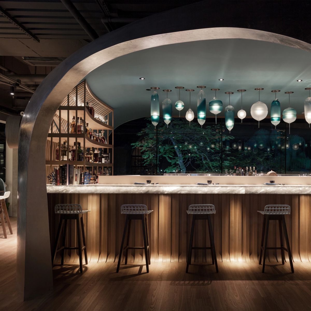 A modern, dimly lit bar with an arched ceiling, marble counter, wooden ribbed facade, and unique blue and white pendant lights. A liquor shelf is visible behind the counter, with a large window looking out to illuminated trees at night.
