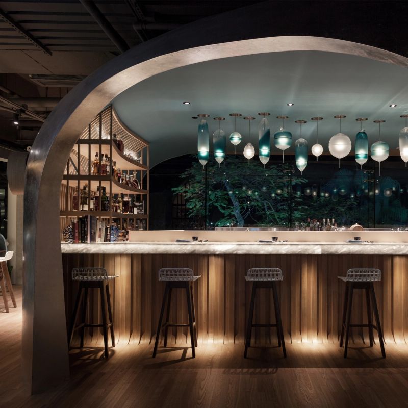 A modern, dimly lit bar with an arched ceiling, marble counter, wooden ribbed facade, and unique blue and white pendant lights. A liquor shelf is visible behind the counter, with a large window looking out to illuminated trees at night.