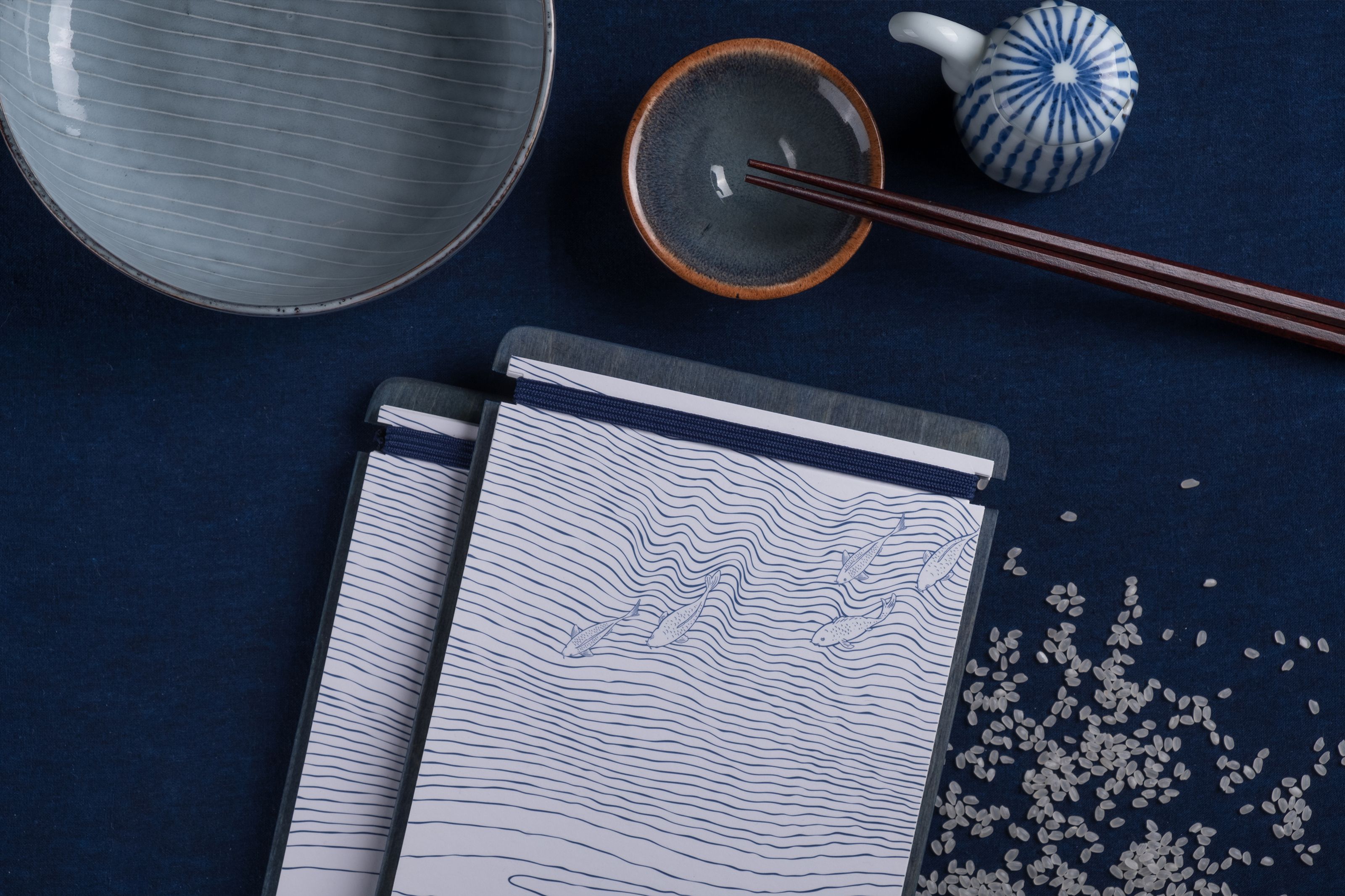 A dark blue table setting with a light blue bowl, chopsticks in a small dish, a decorative ceramic piece, scattered rice, and two menus featuring a blue wave and fish design.