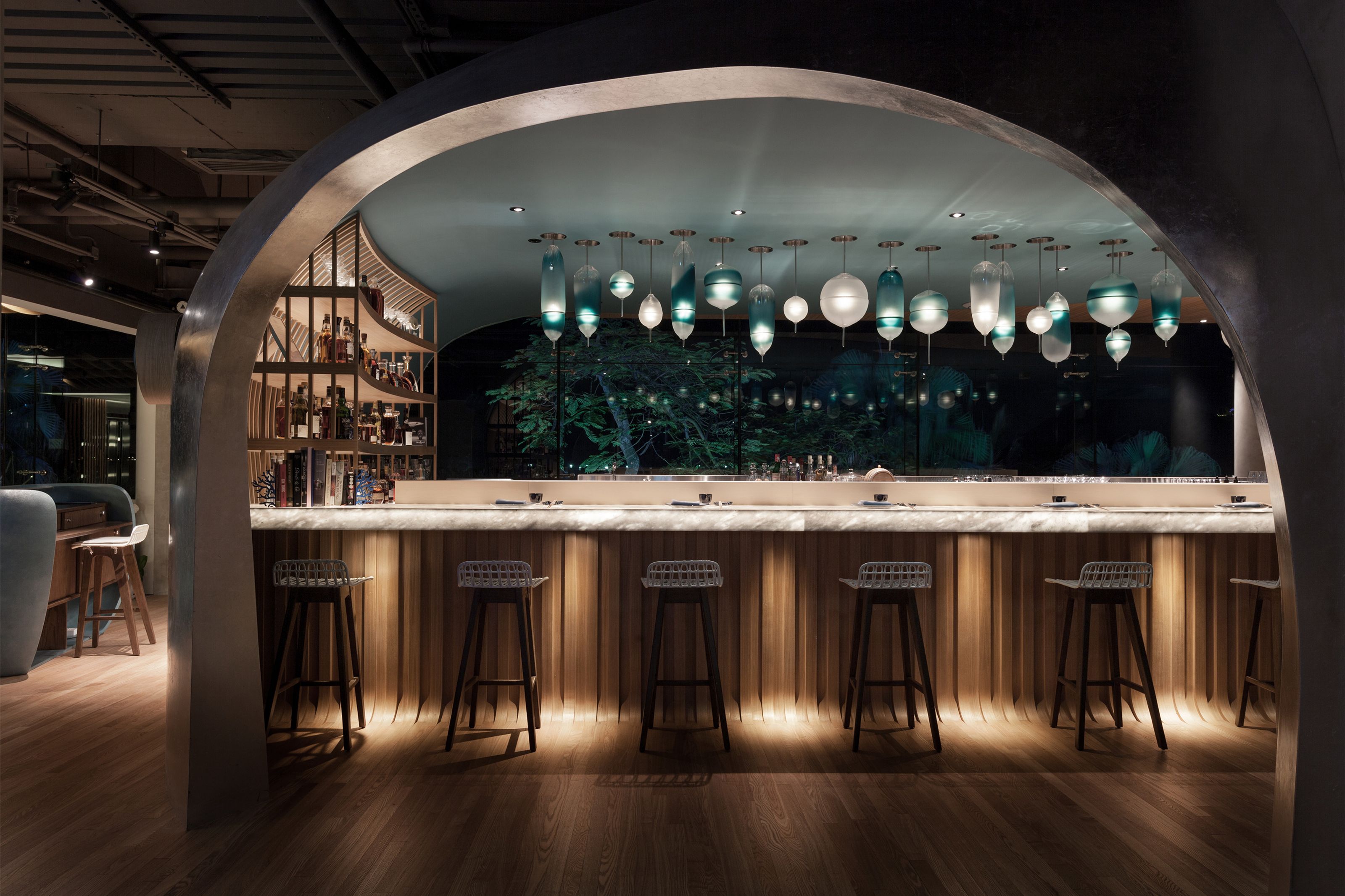 A chic bar with a white marble counter, wooden facade, and stools, under anway archway, illuminated by unique blue and white pendant lights, with a night view of trees.