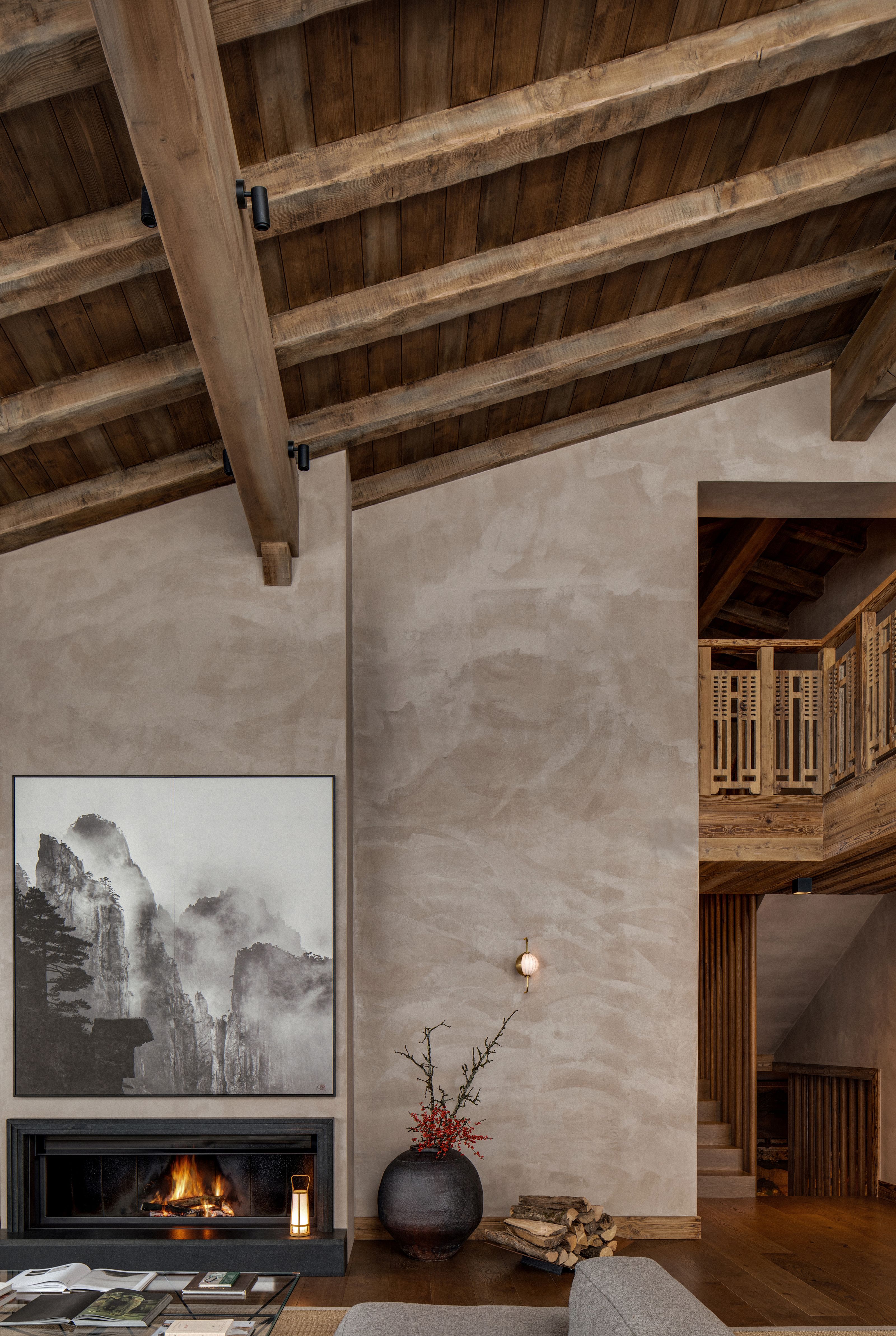 A warm interior with a pitched wooden ceiling, a lit fireplace under a misty mountain artwork, and a wooden staircase.