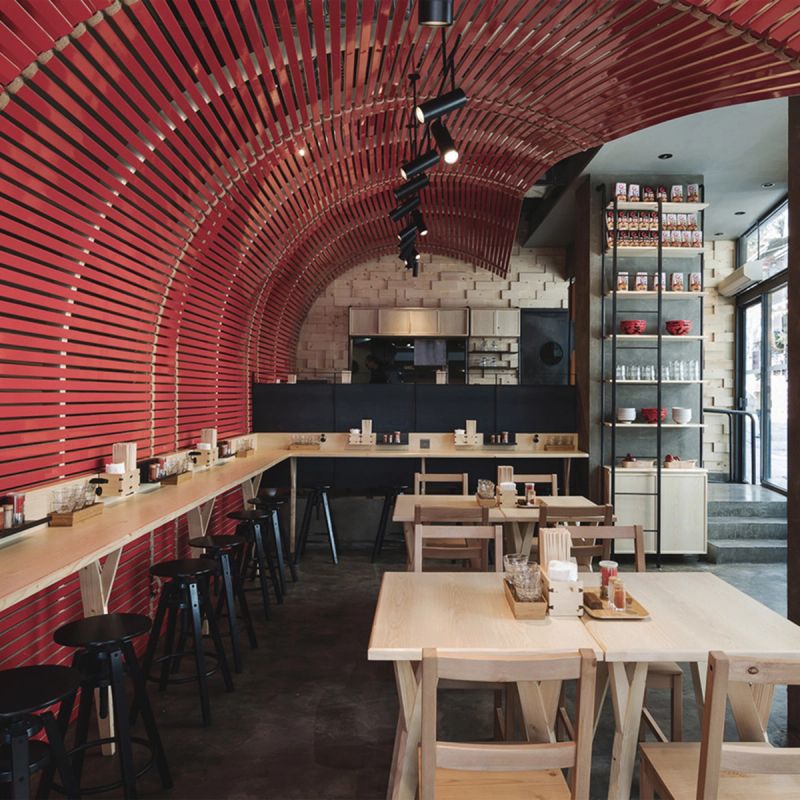 Modern restaurant interior featuring a red slatted arched ceiling and light wood furniture.