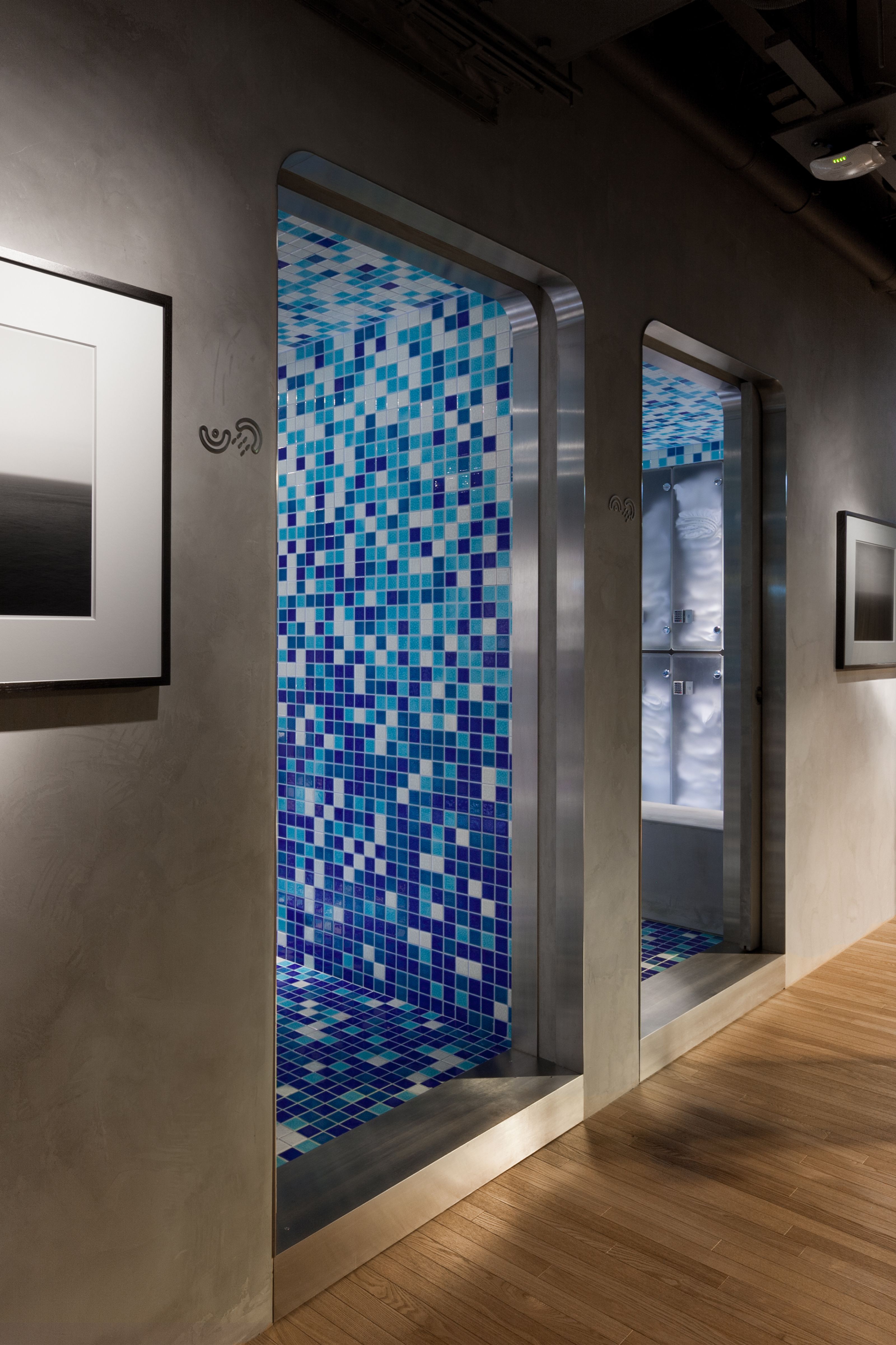 A modern hallway with gray walls and a wooden floor, featuring two arched entrances leading to rooms tiled with blue and white mosaic. A framed artwork hangs on the left.