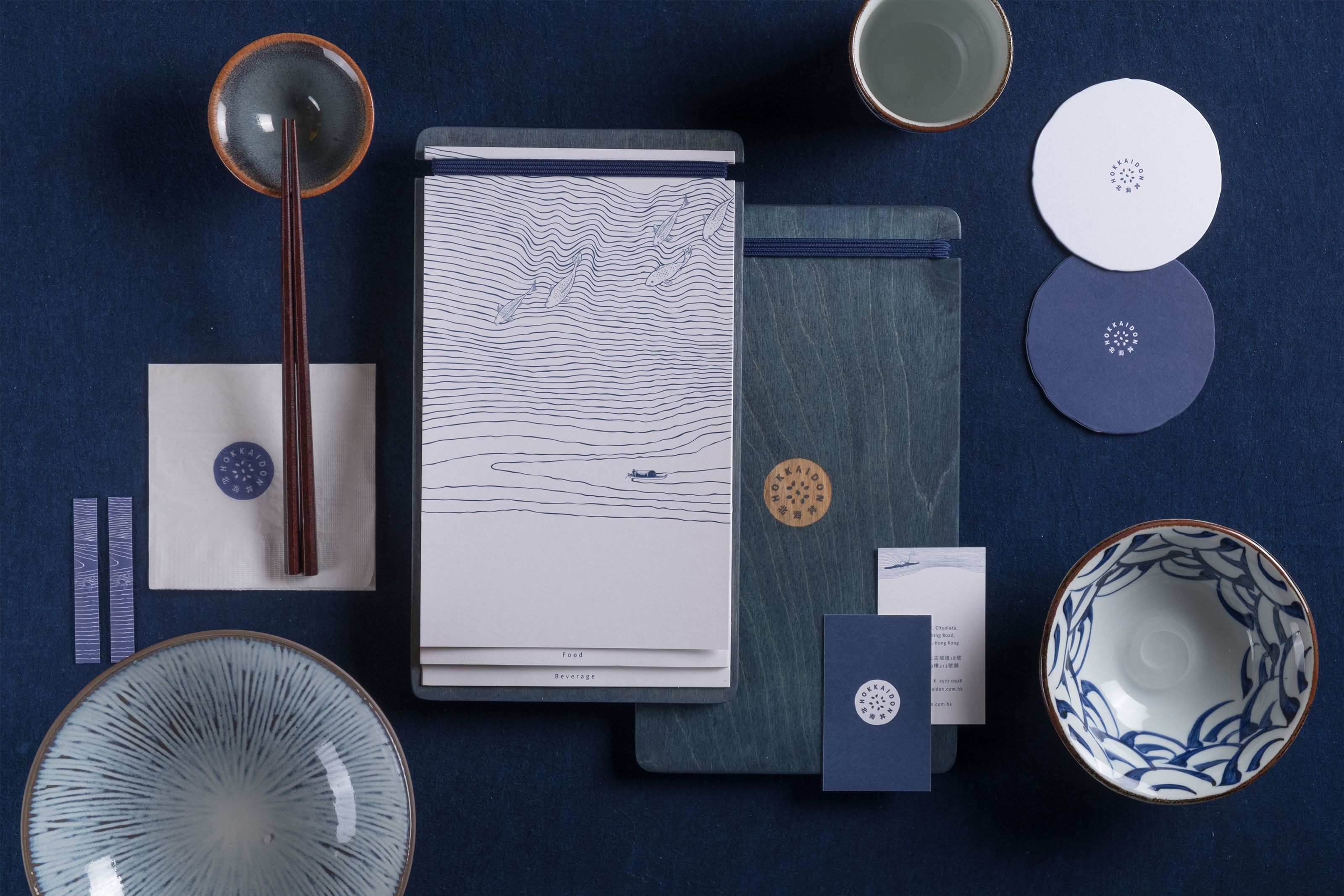 A flat lay of blue and white restaurant branding and tableware, including a menu with a wave pattern, ceramic bowls, chopsticks, and coasters, on a dark blue background.