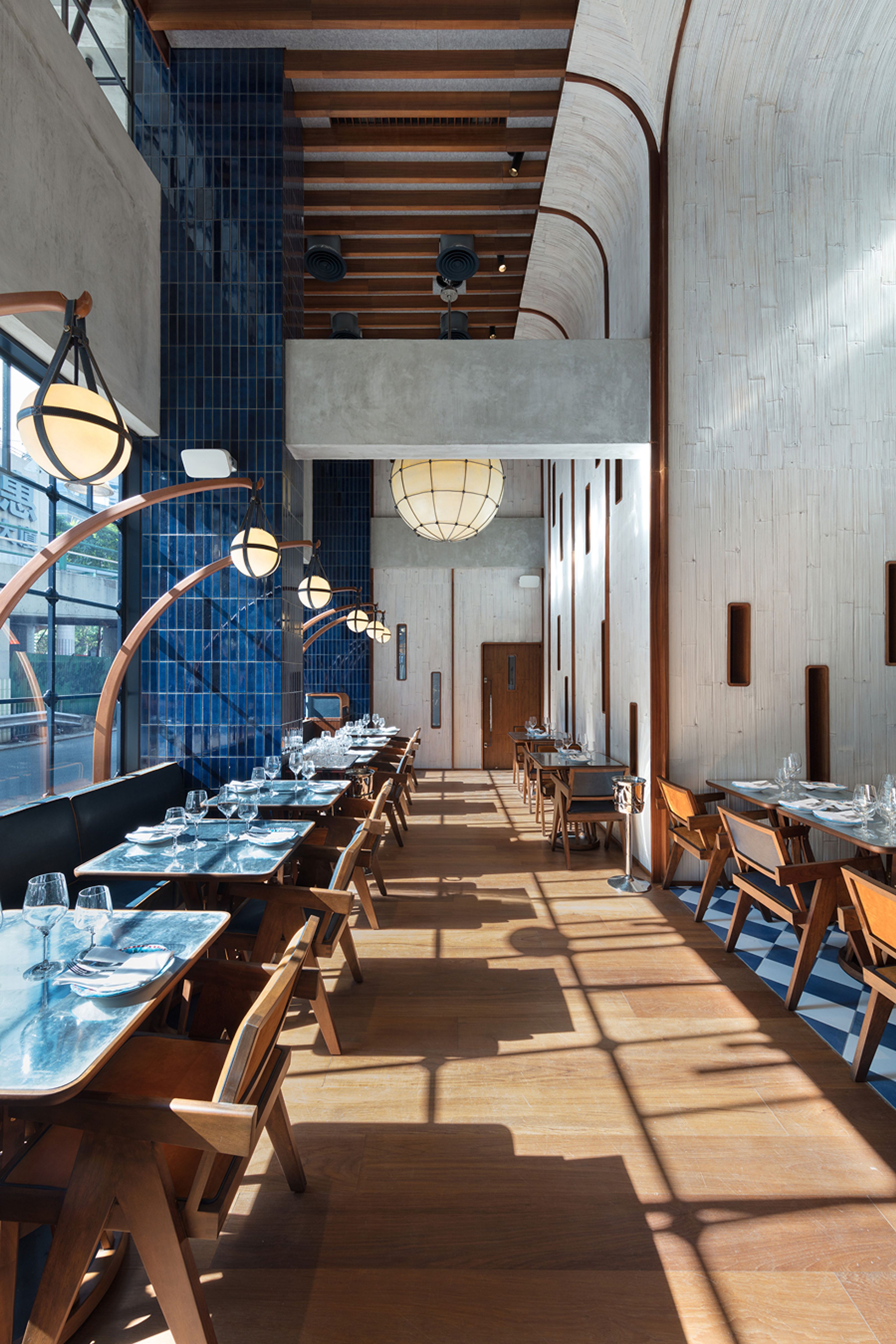 An empty, modern restaurant dining room with blue tiled walls, light wooden floors, spherical pendant lights, and sunlight creating patterned shadows.
