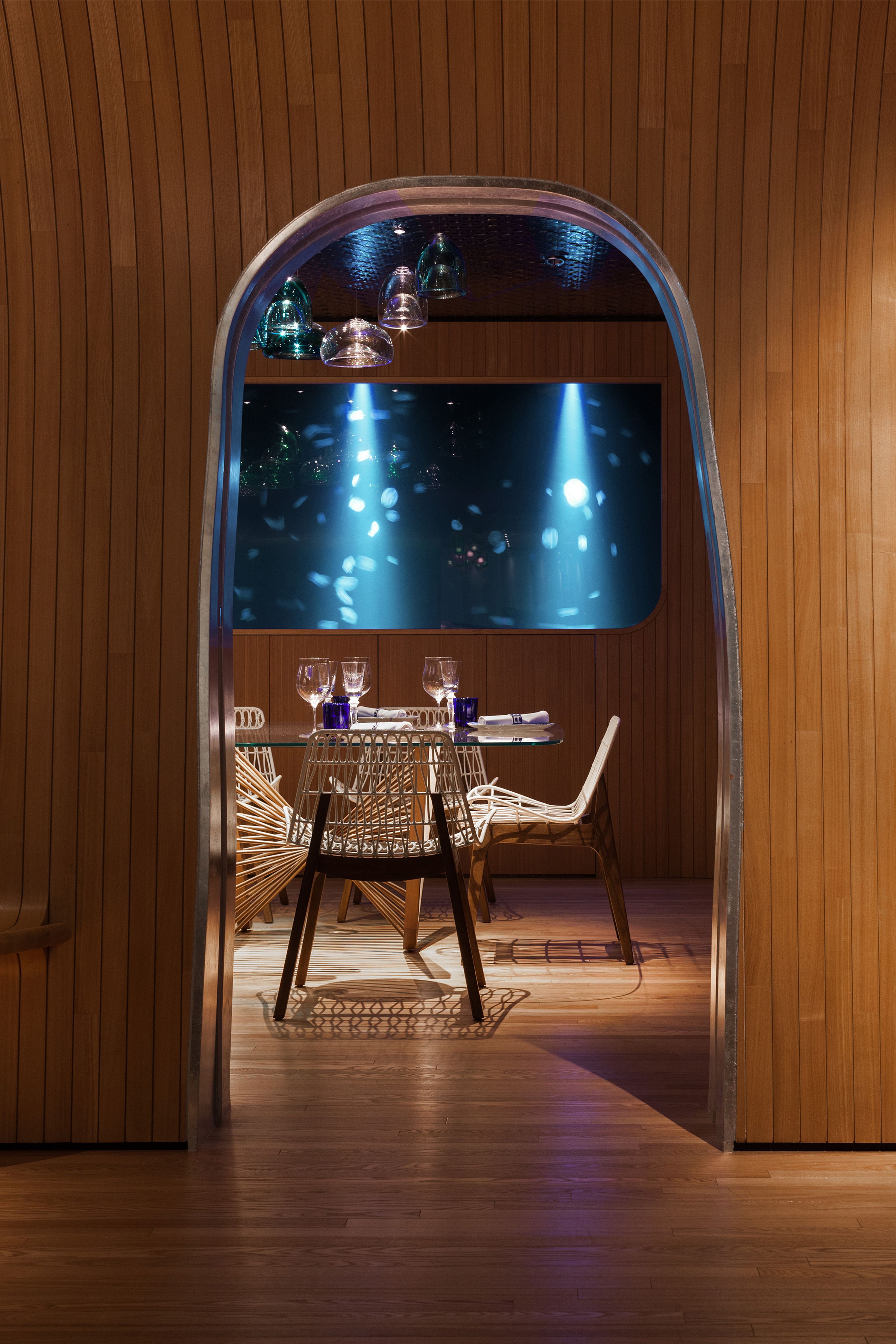 A modern dining room with a glass table and unique chairs, viewed through a wooden archway, facing a large blue-lit aquarium displaying jellyfish.