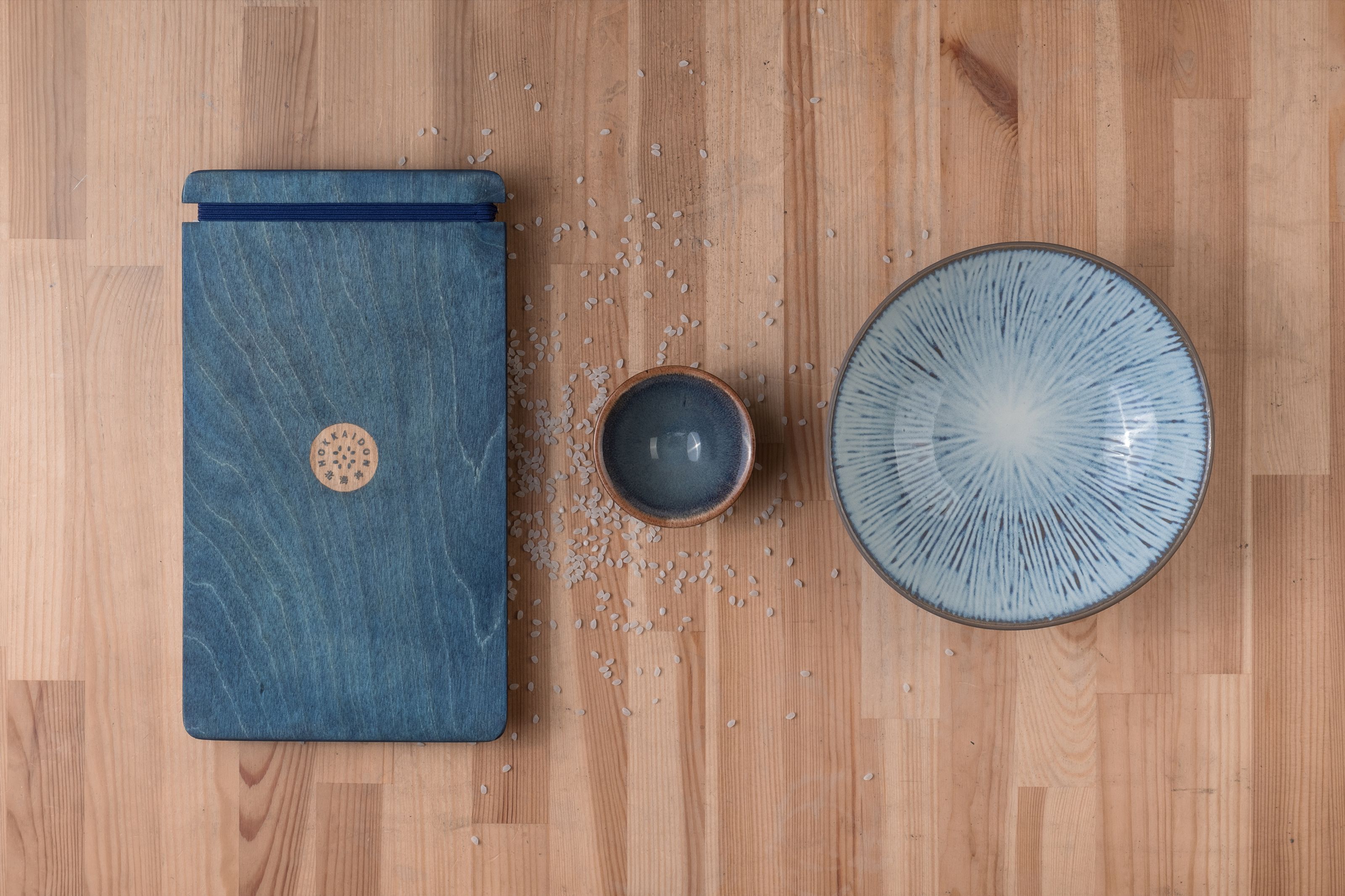 A blue wooden menu, a small blue cup, and a patterned blue bowl with scattered rice grains on a light wooden table.