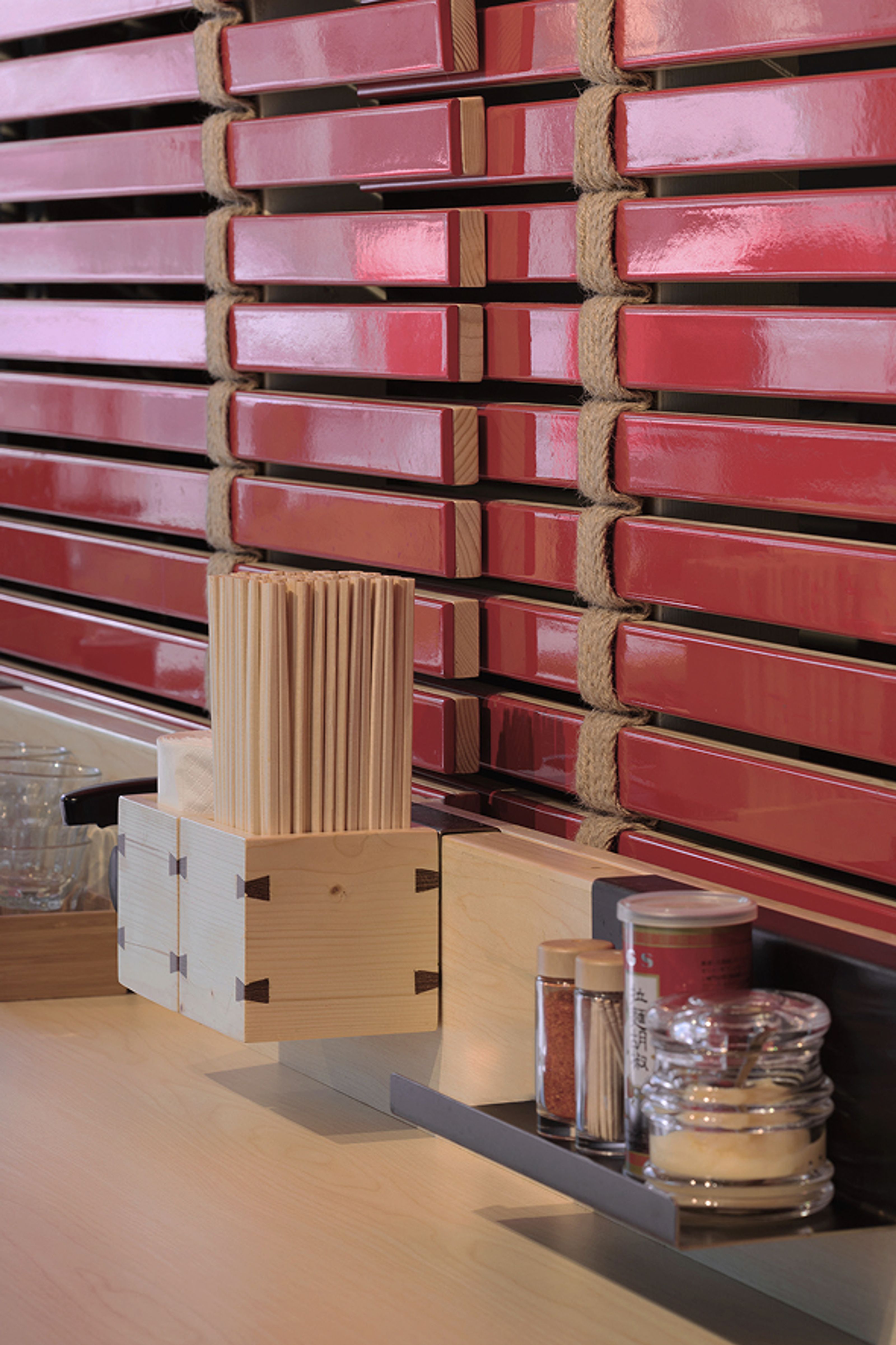 A light wooden counter holds a box of chopsticks and various condiments, set against a wall of horizontal red lacquered slats bound by rope.