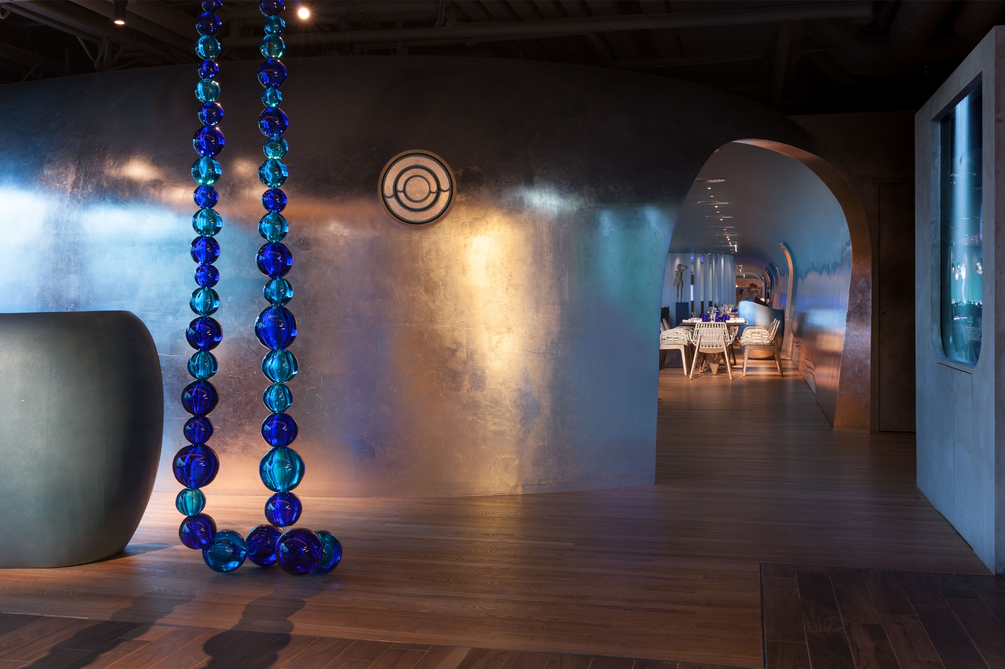 Modern interior with a curved metallic wall, decorative blue and teal glass bead strands, and an arched doorway to a dining room.