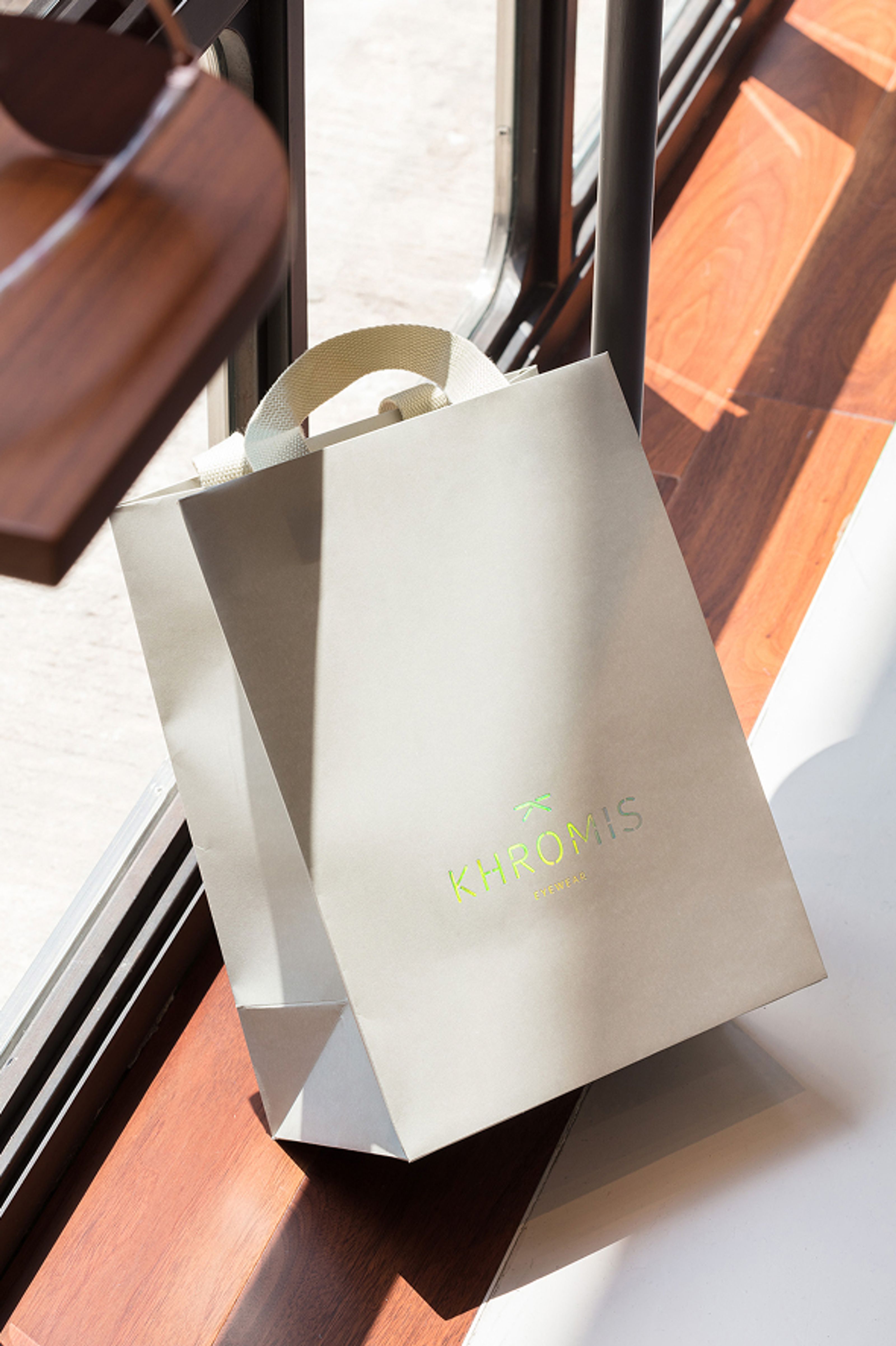 A light gray shopping bag with a neon green "KHROMIS EYEWEAR" logo on a sunlit wooden floor.