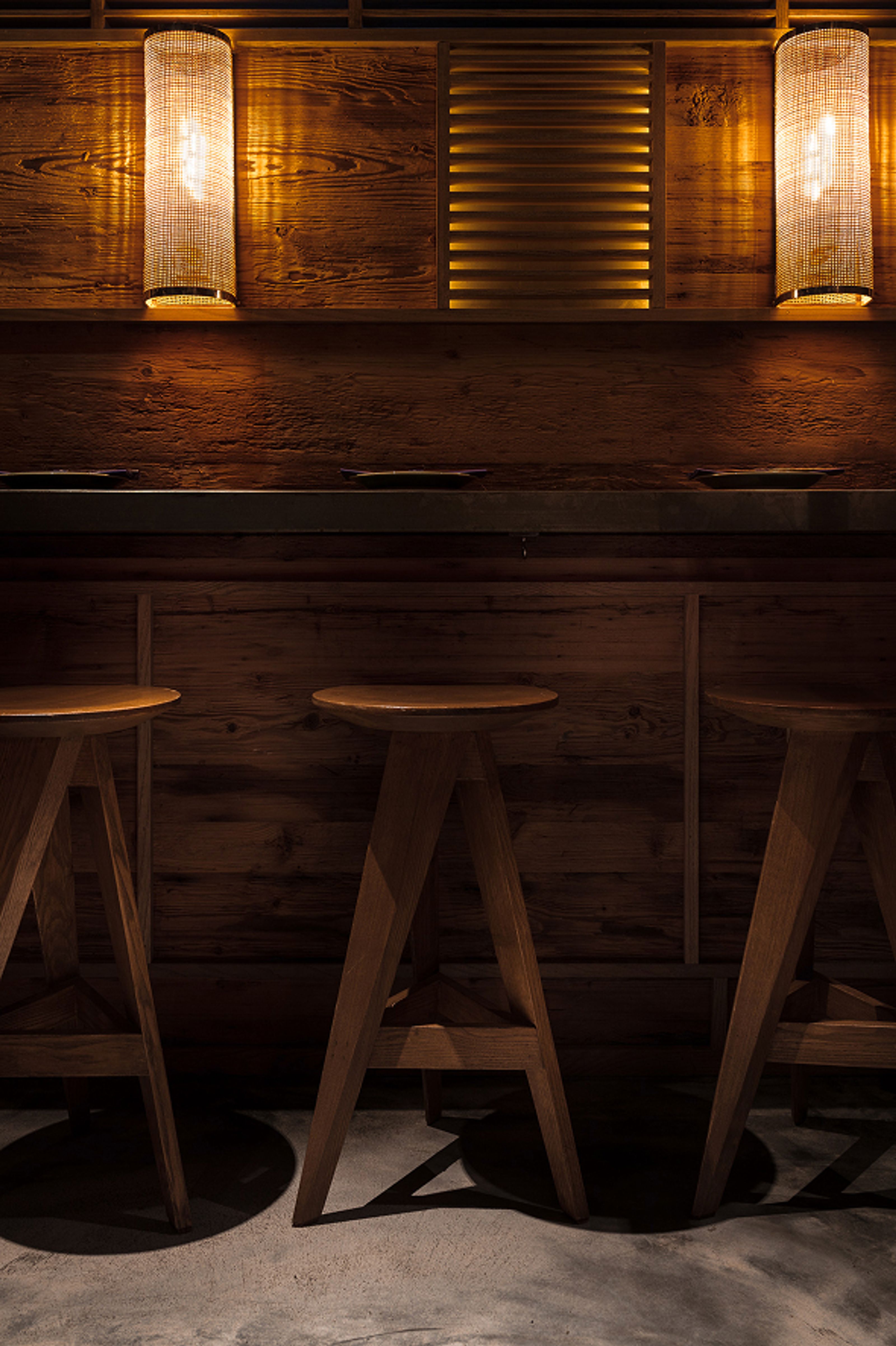 Warmly lit wooden bar with three stools and two wall sconces.