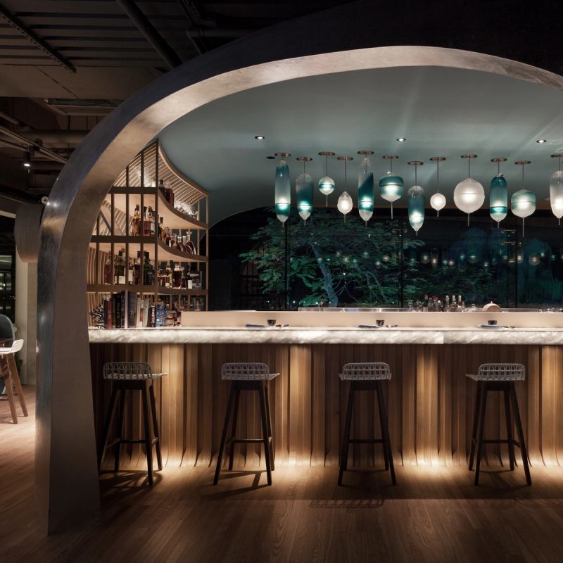 A modern, dimly lit bar with an arched ceiling, marble counter, wooden ribbed facade, and unique blue and white pendant lights. A liquor shelf is visible behind the counter, with a large window looking out to illuminated trees at night.