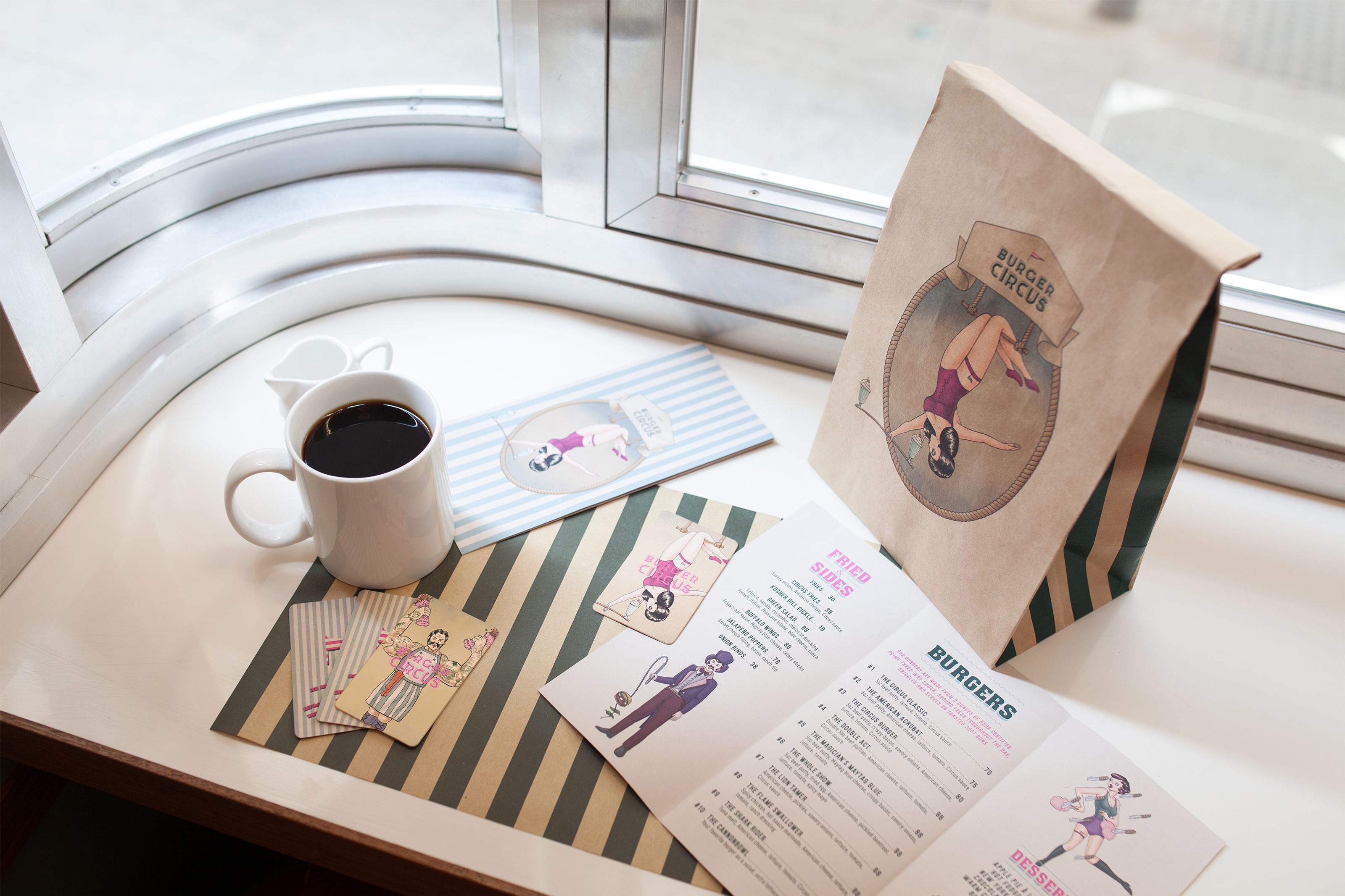A Burger Circus branded paper bag, open menu, coffee, and circus-themed pin-up illustrations on a table by a window.