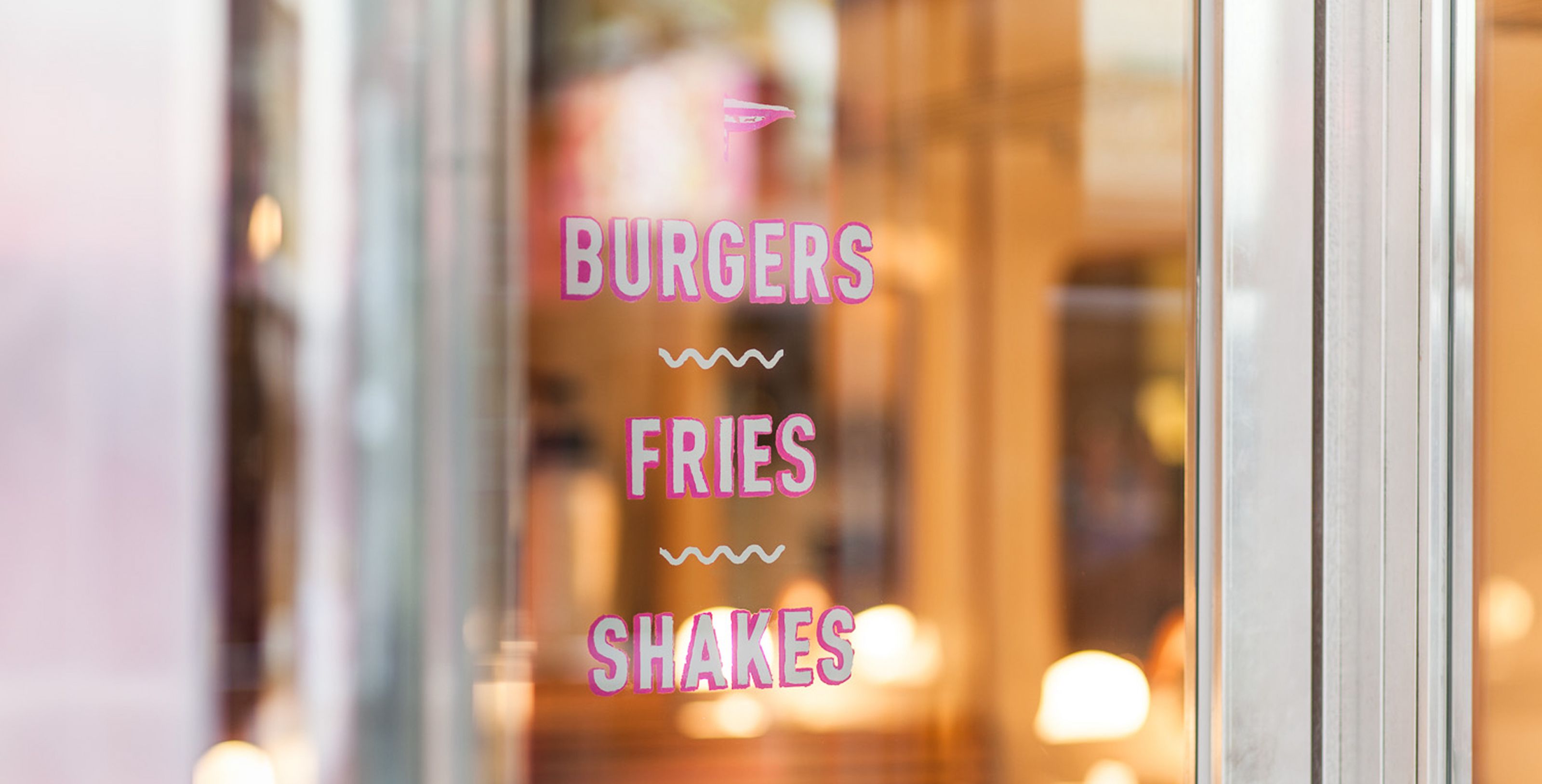 Pink text on a glass door advertising "BURGERS", "FRIES", "SHAKES".