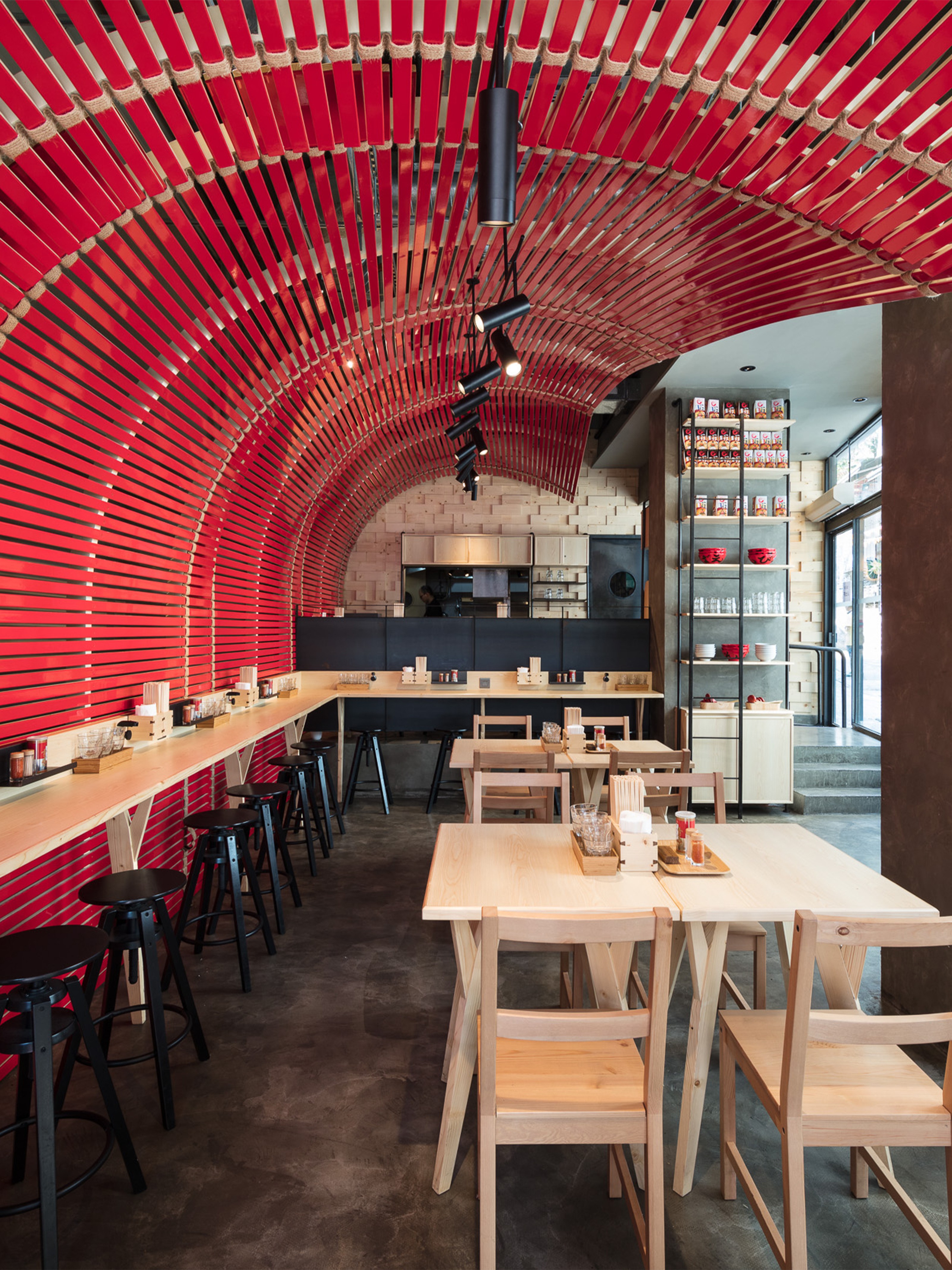 Interior of a modern restaurant with a vibrant red slatted arched ceiling and wall, light wood furniture, and concrete flooring.