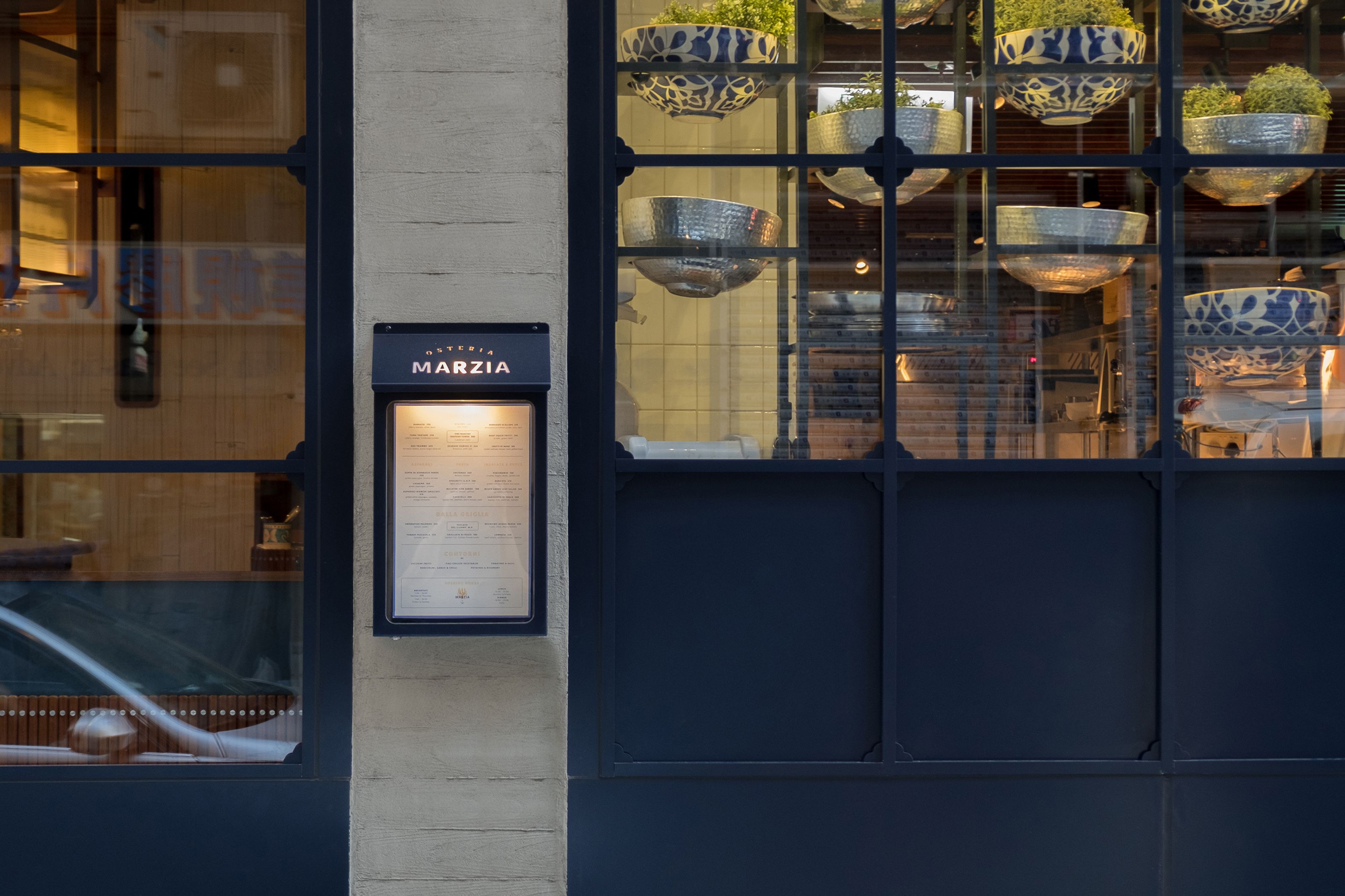 Osteria Marzia exterior with a lit menu board, large windows, and interior decorative bowls.