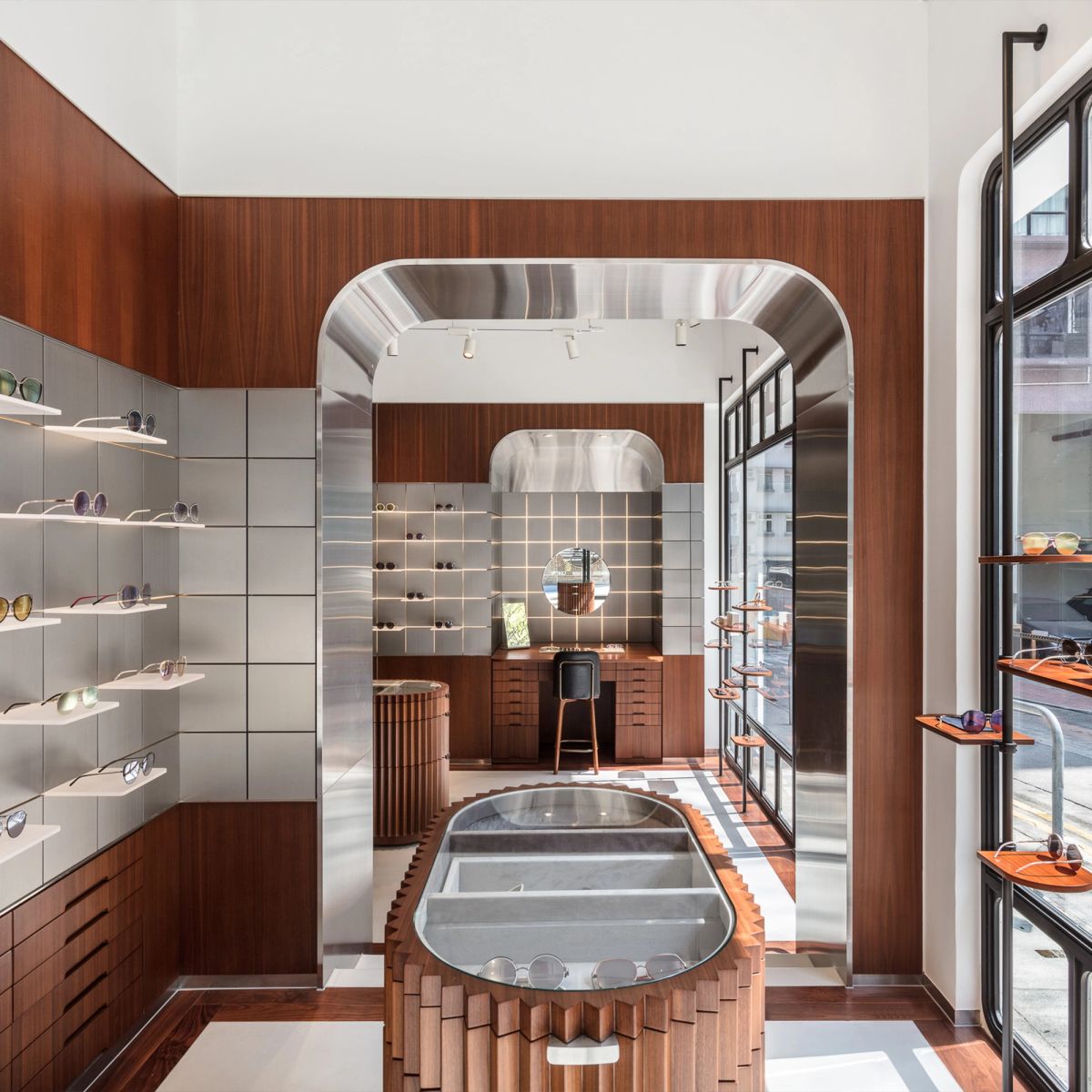 A modern eyewear store featuring warm wood paneling, gray tiled walls with glasses, an arched metallic entrance, and a central oval wooden display.