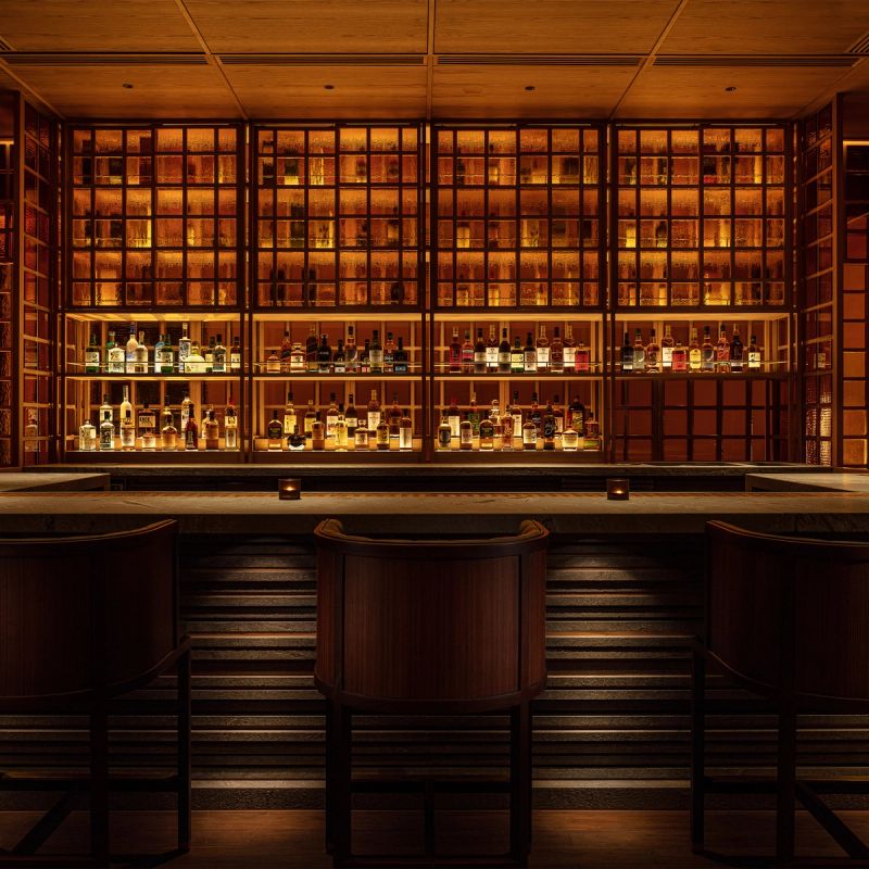 A warmly lit bar with shelves of liquor bottles and two bar stools.