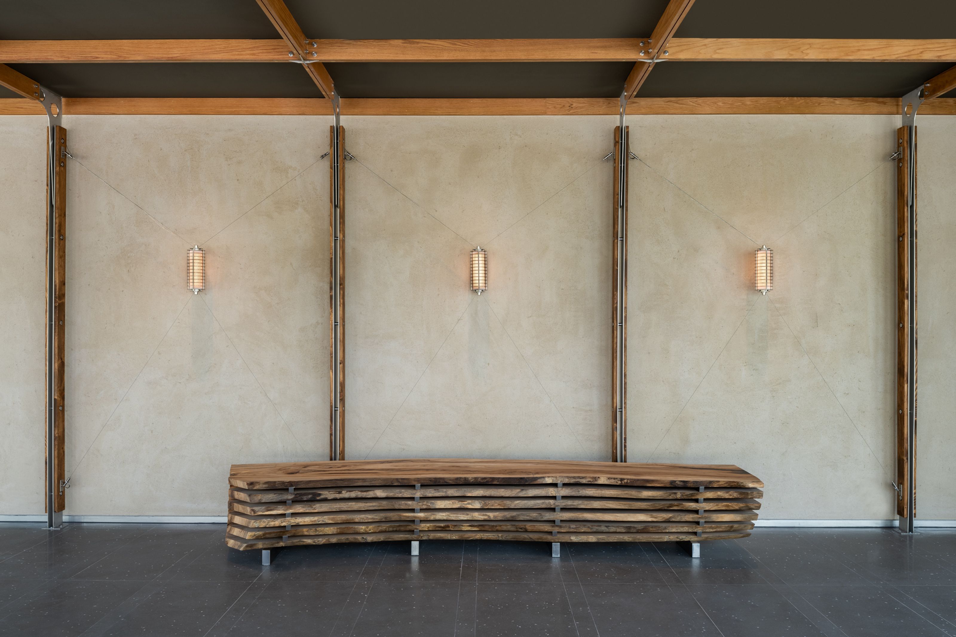 A minimalist room with a long, stacked live-edge wood bench against a light concrete wall, flanked by three glowing cylindrical sconces and vertical wooden beams, under a beamed ceiling.
