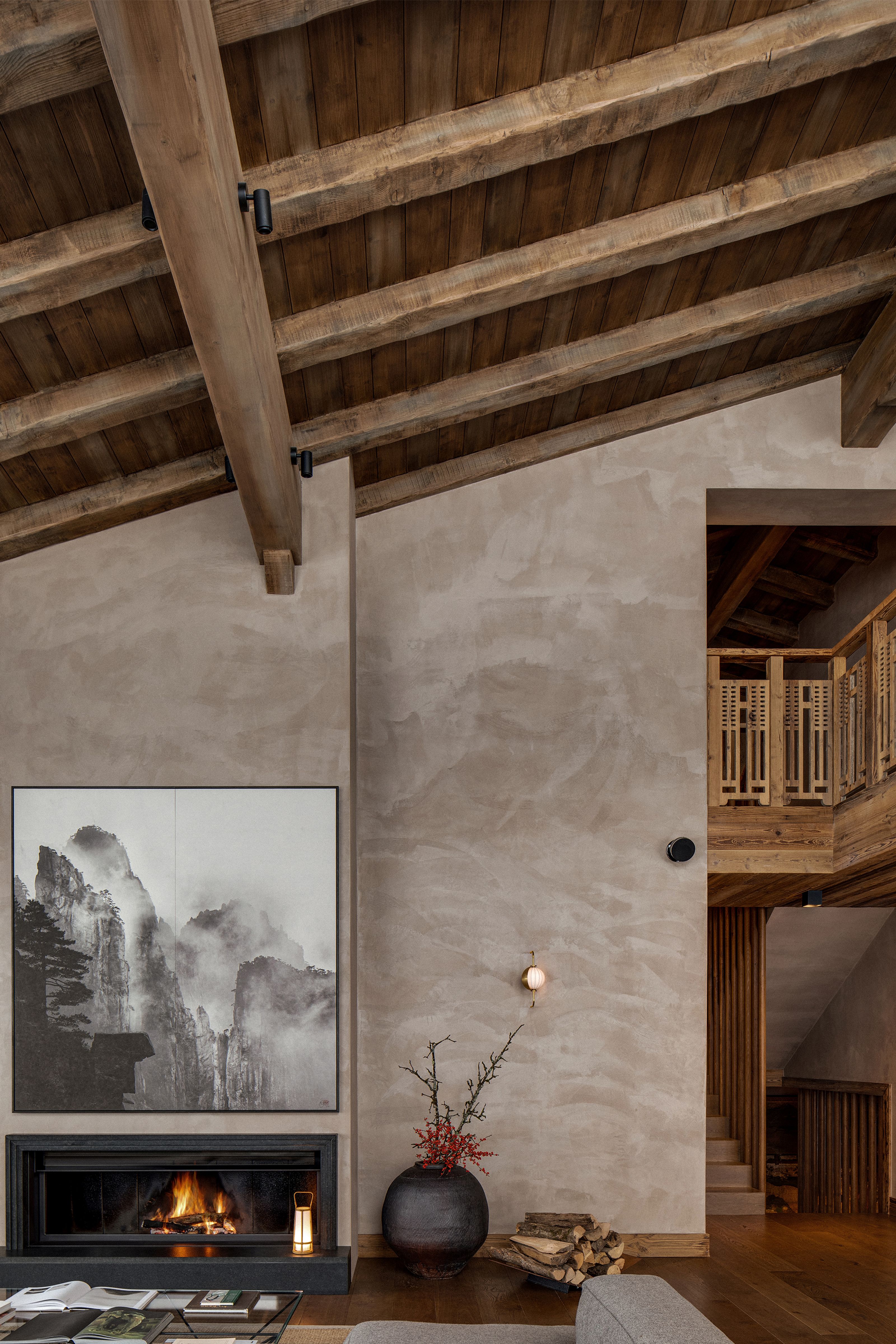 A rustic-modern living room with a vaulted wooden ceiling, a fireplace beneath a large mountain artwork, and a wooden staircase.