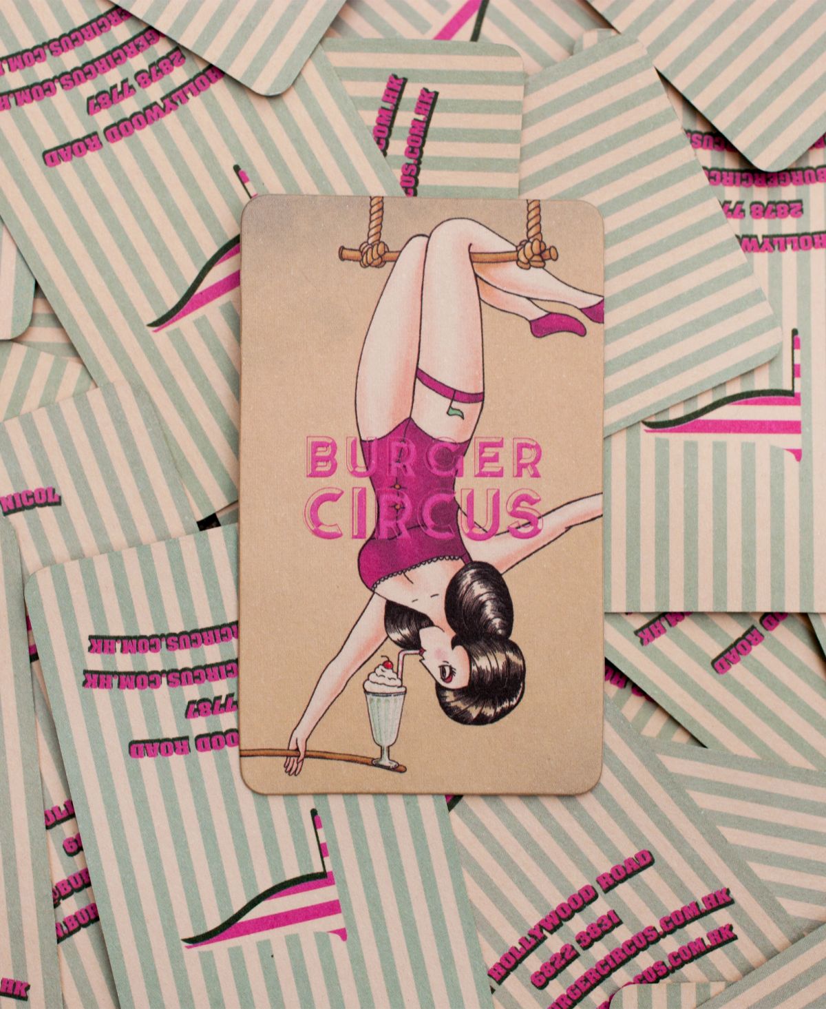 A Burger Circus business card featuring an upside-down pin-up trapeze artist drinking a milkshake, surrounded by other striped cards showing the restaurant's details.