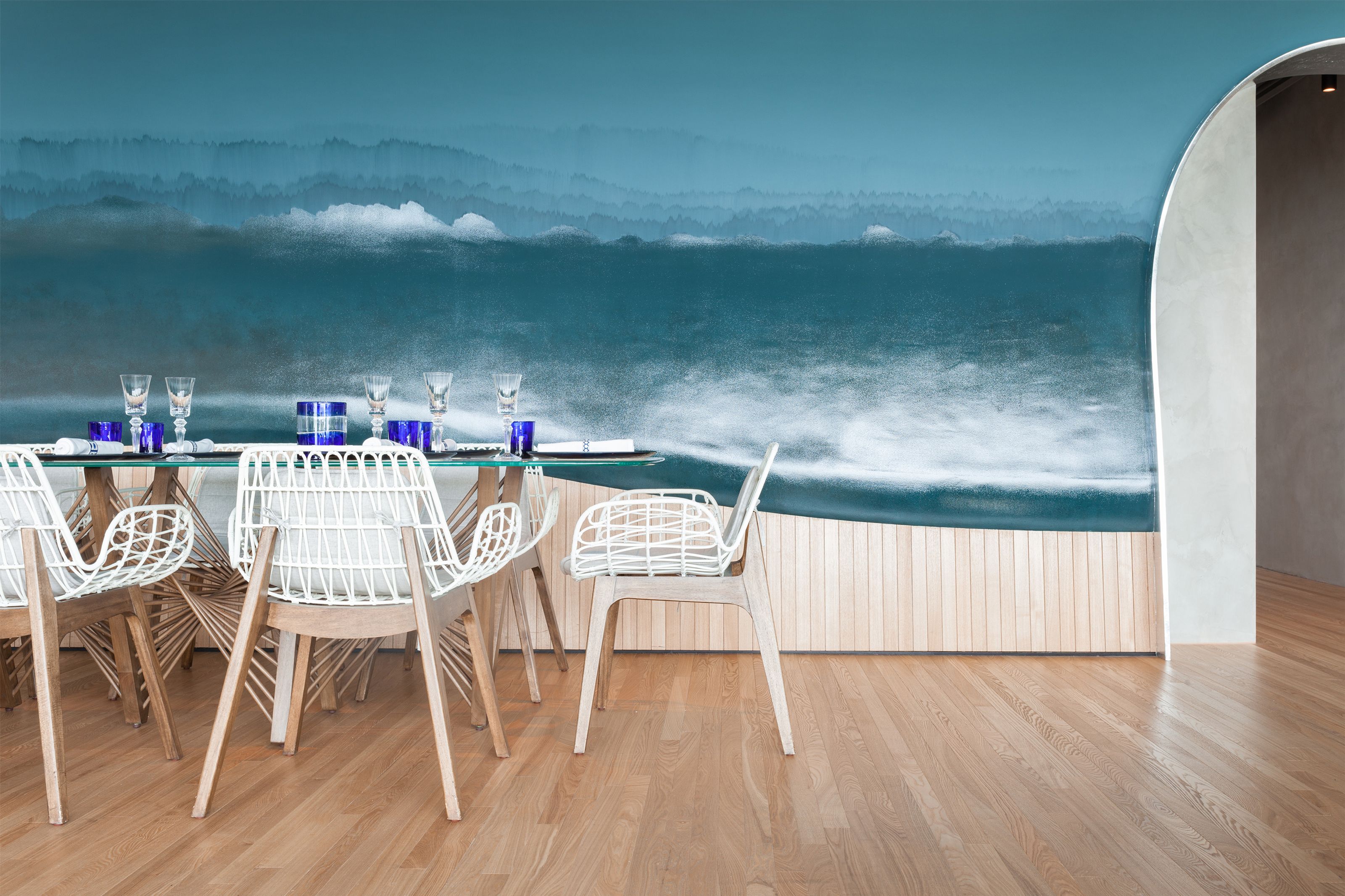 Restaurant interior with a floor-to-ceiling ocean wave mural behind a glass table set with blue glasses and surrounded by white chairs.