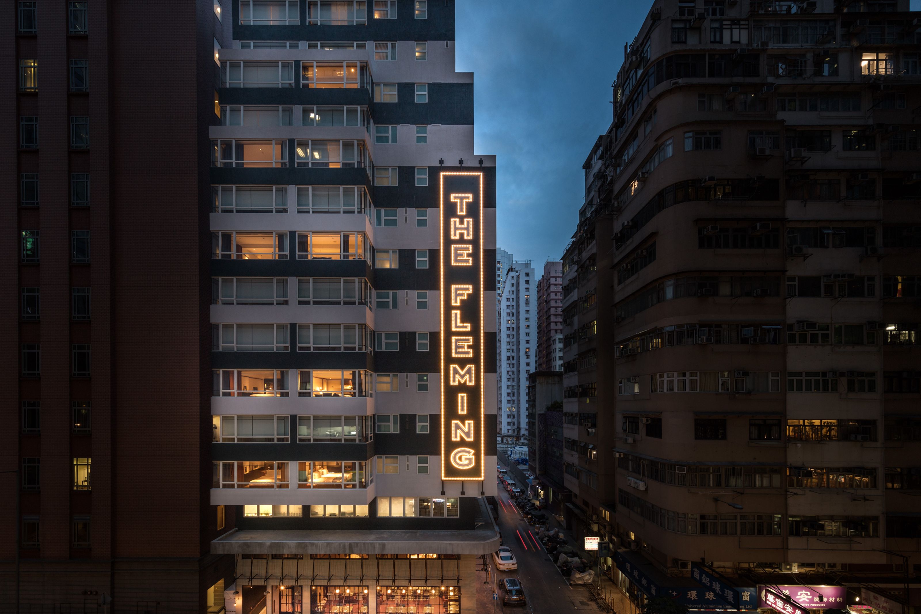 Neon sign "THE FLEMING" on a modern building in a city at dusk, with other buildings and a street below.