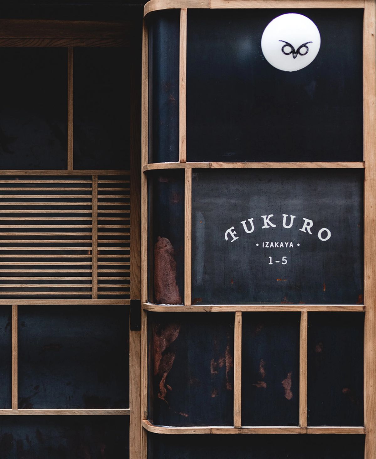 Dark-paneled building entrance with light wood grid framing, a white owl logo, and "FUKURO IZAKAYA 1-5" text.