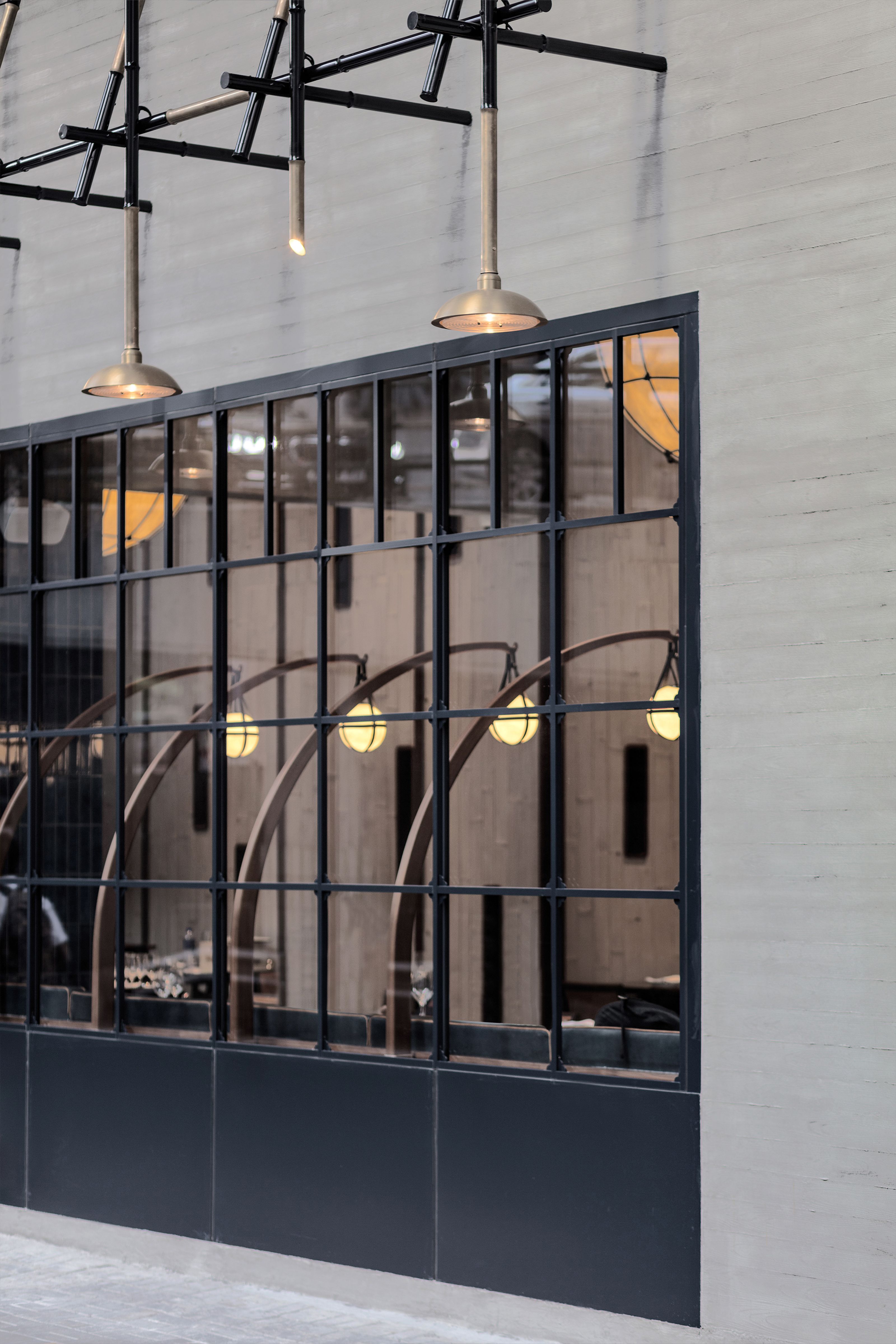 Industrial window with black grid framing unique curved lamps inside, brass pendants above, and a textured concrete wall.