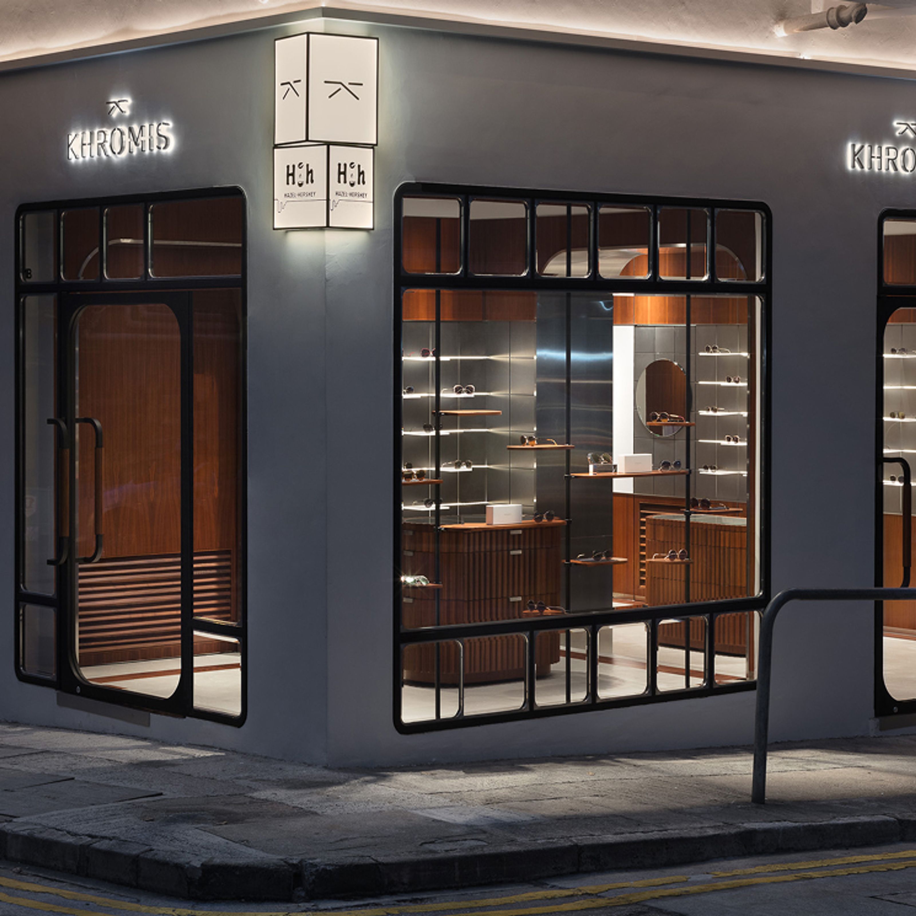 Corner view of a modern eyeglasses store at night, with illuminated "KHROMIS" and "H&H" signs and windows displaying eyewear.