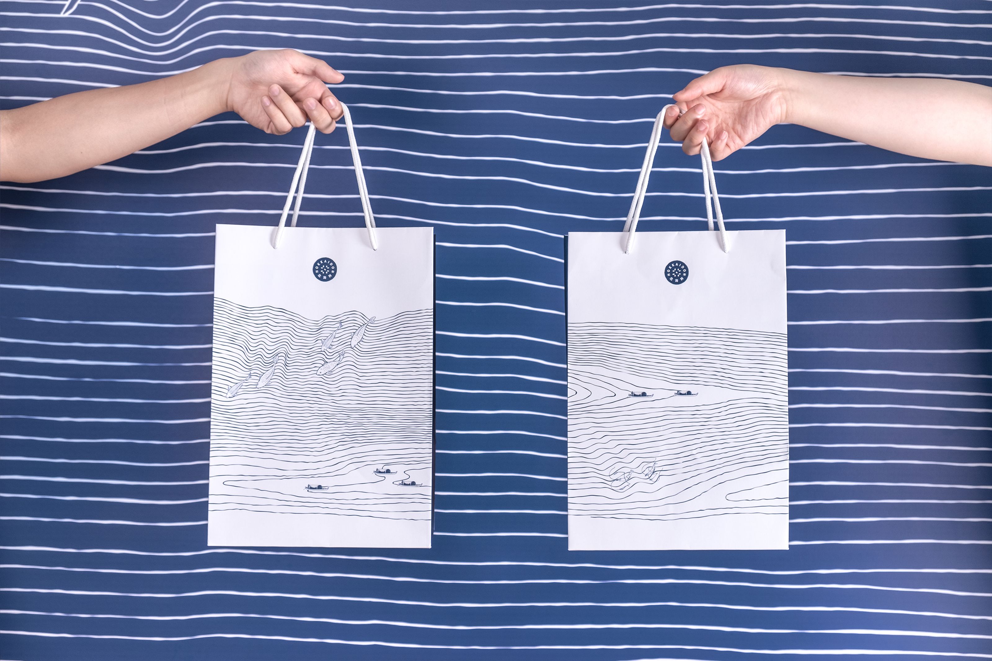 Two hands hold white gift bags with blue line-art landscapes against a blue striped background.