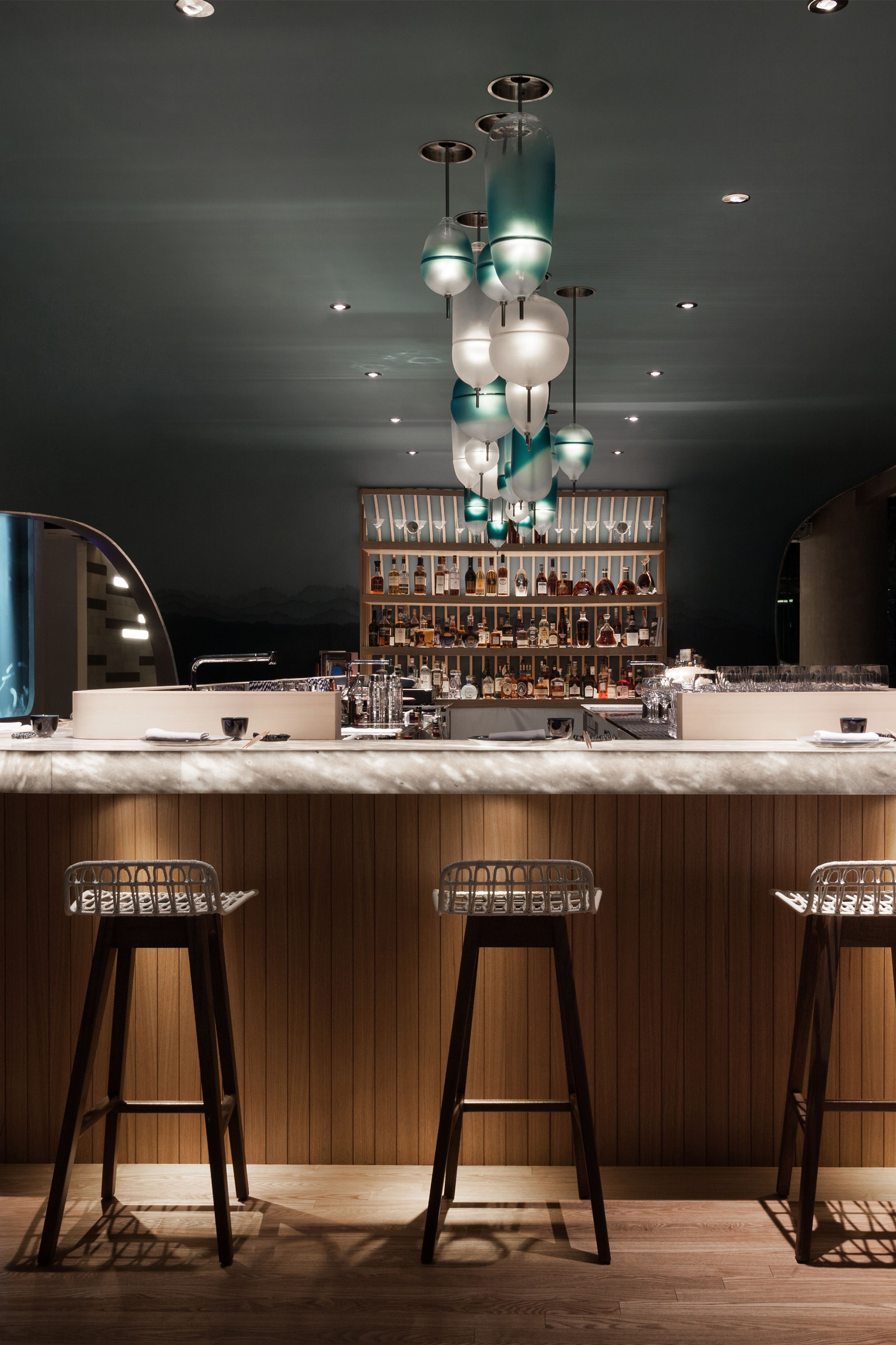 A stylish bar interior with a marble counter, wood paneling, three bar stools, a liquor shelf, and a cascade of frosted white and teal glass pendant lights.