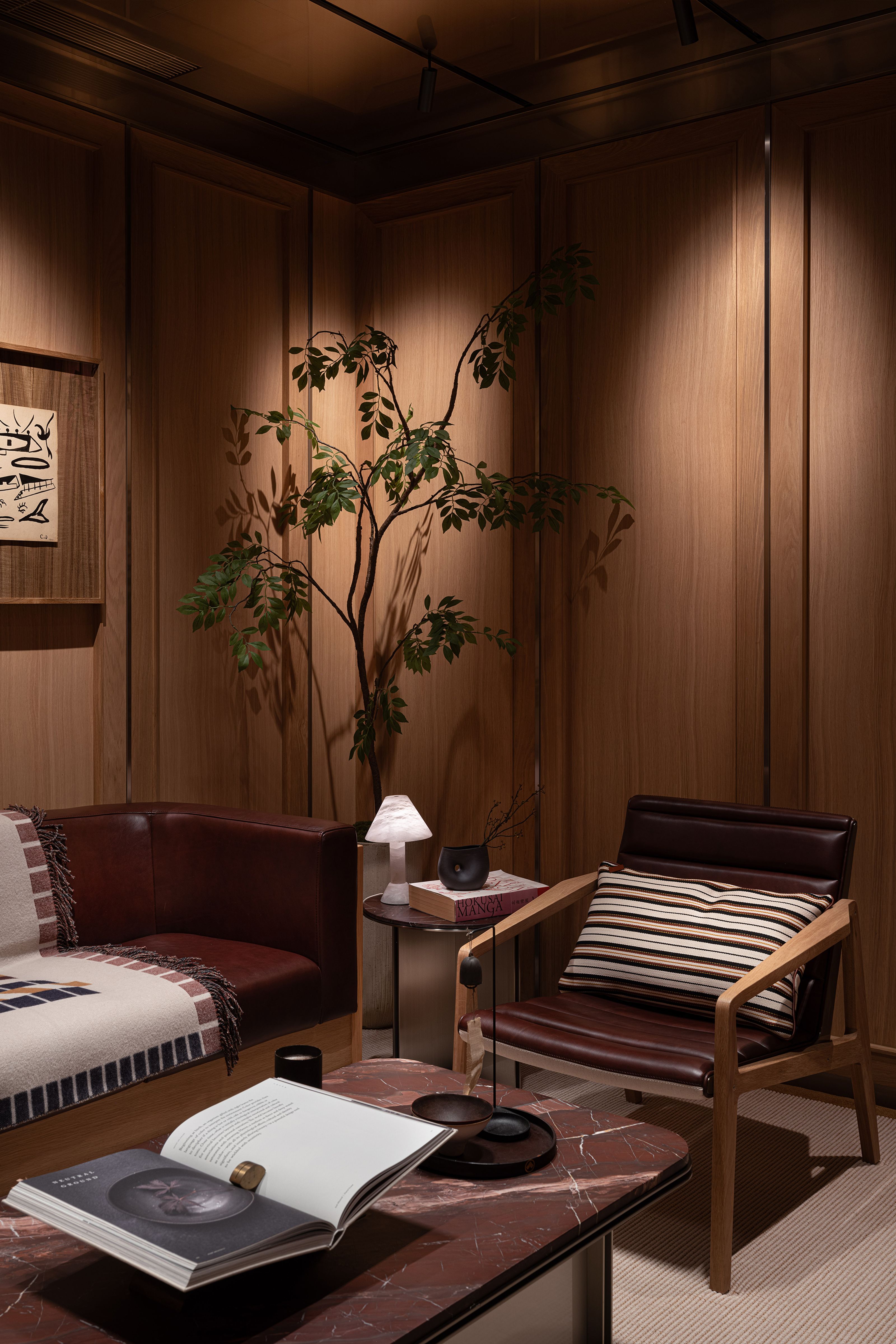 A warm, wood-paneled room with brown leather furniture, a reddish marble coffee table, and a tall plant casting shadows.