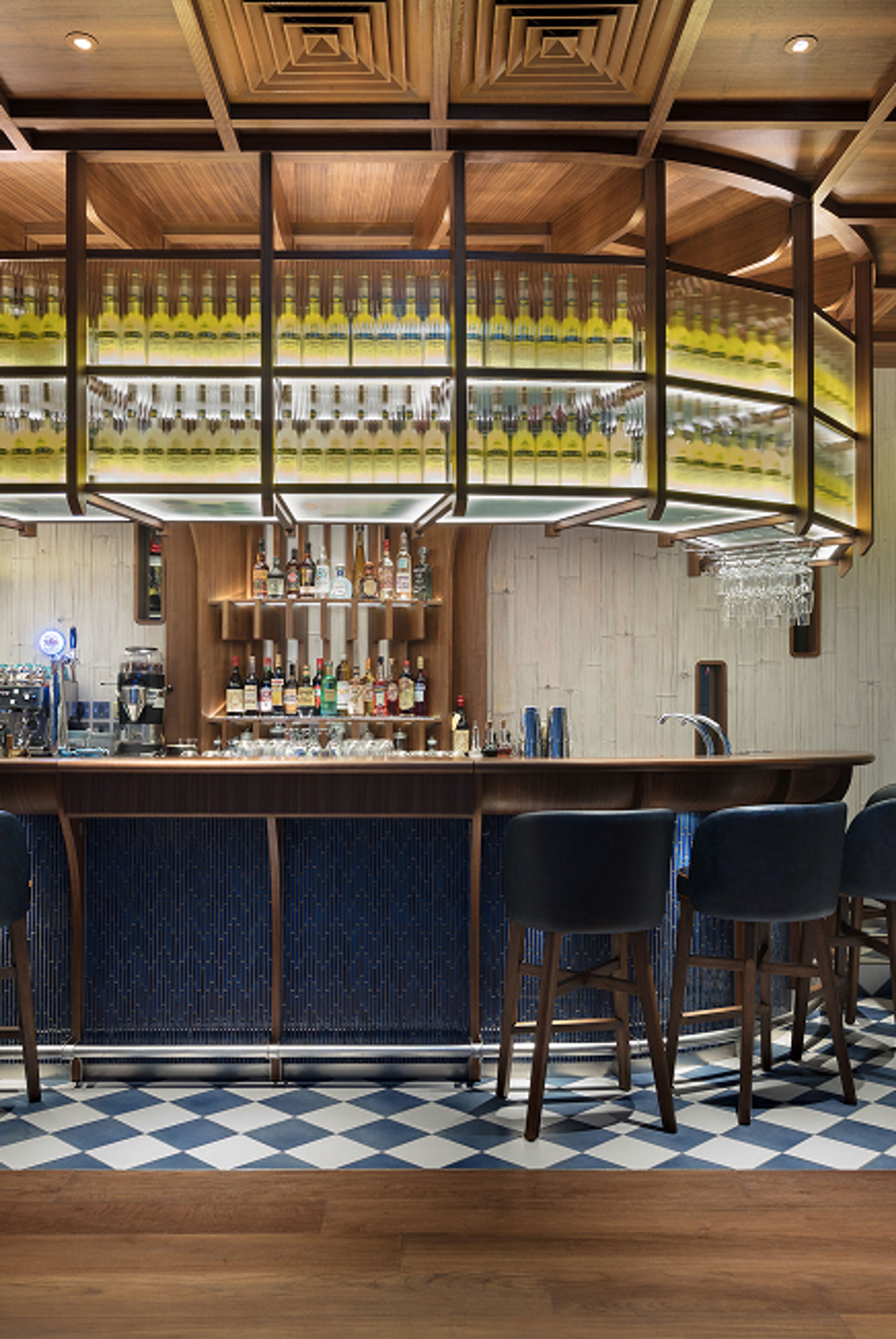 A modern bar interior with a wooden coffered ceiling, a dark wood bar counter with blue paneling, backlit shelves displaying numerous bottles, and a blue and white checkered floor.