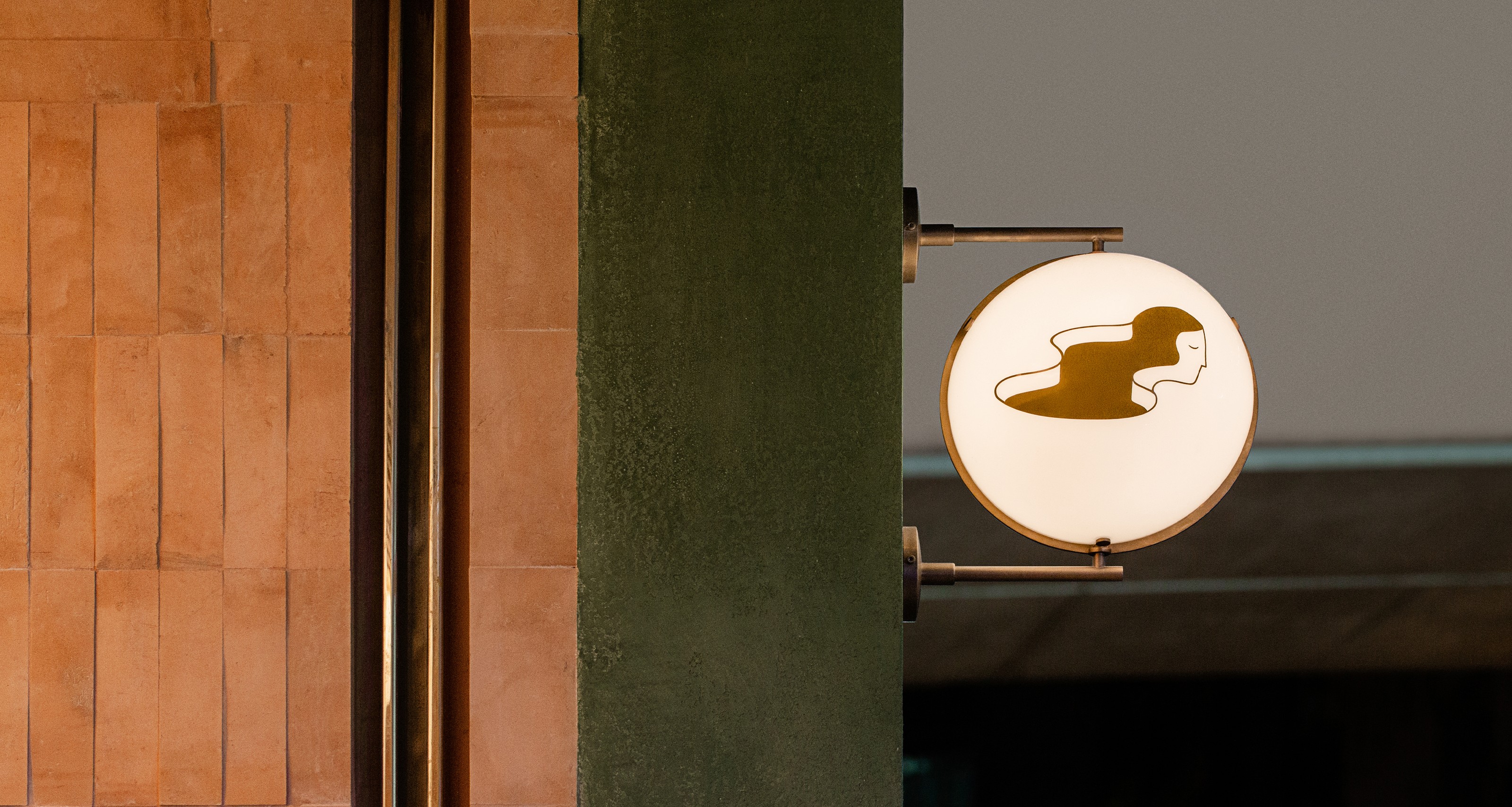 A round illuminated sign with a brown silhouette of a stylized figure, on a green wall next to a terracotta tiled wall.