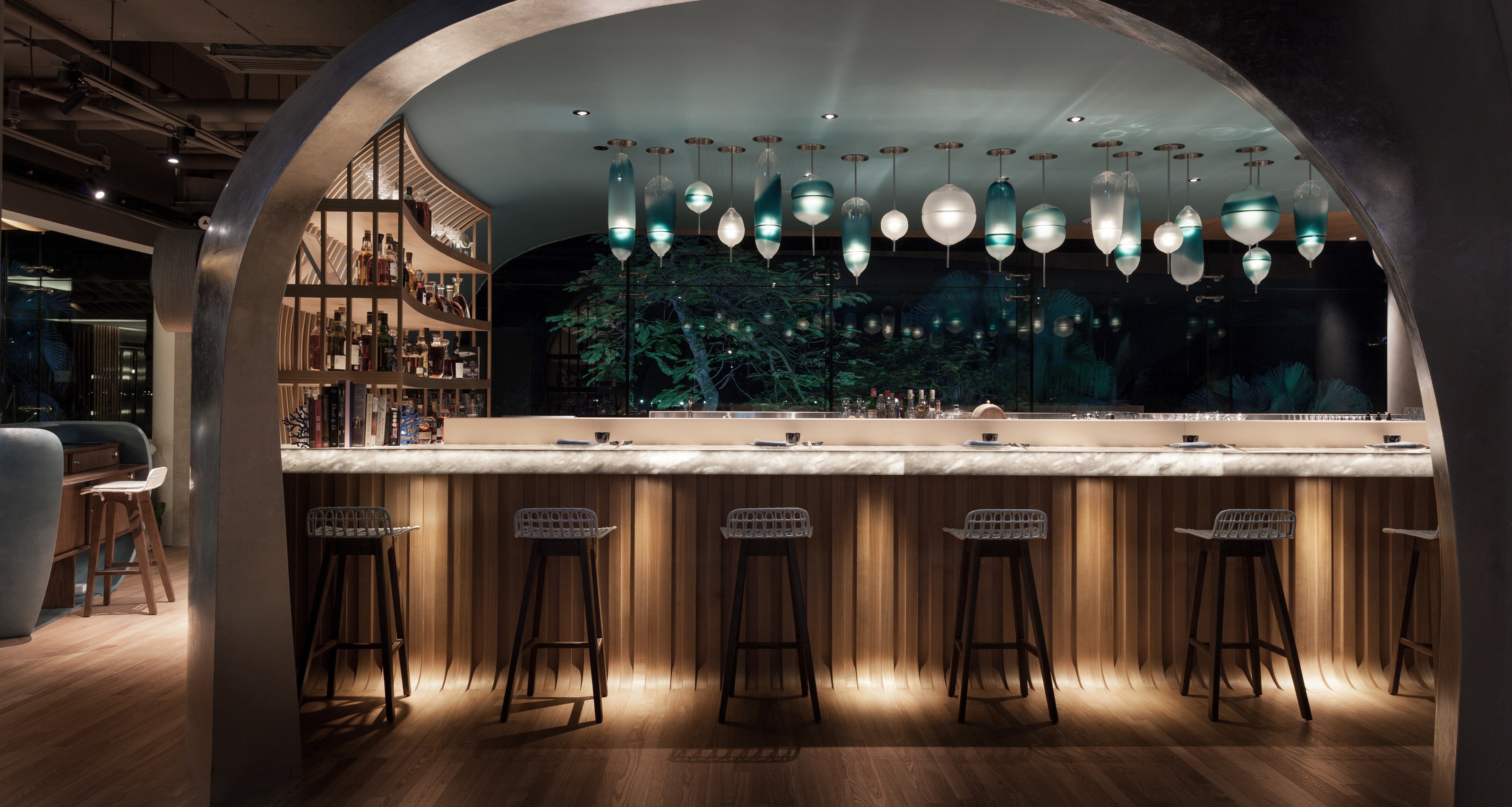 A modern bar with a marble counter, wood paneling, unique glass pendant lights, and a dark outdoor view of trees.
