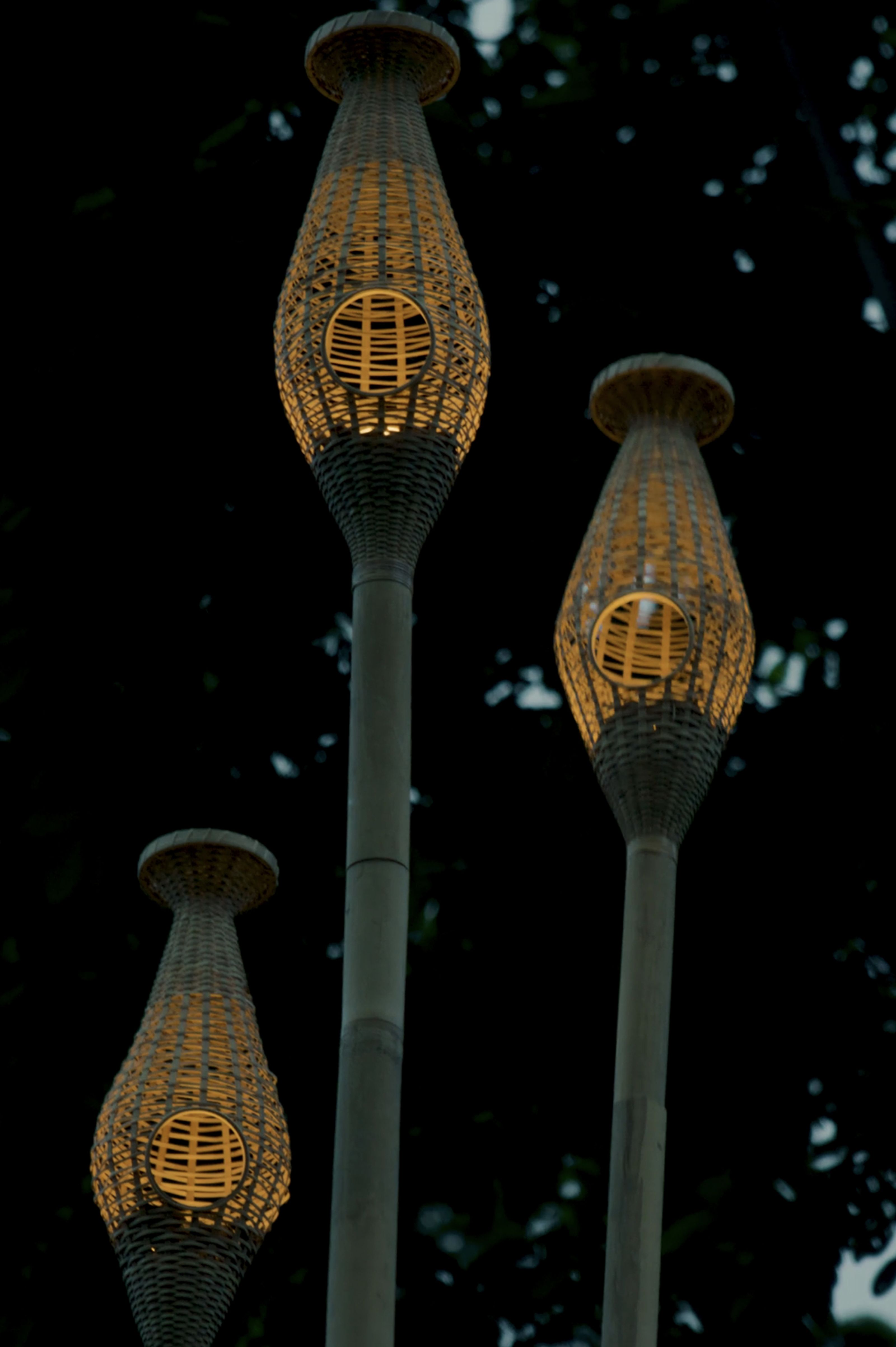 Three illuminated woven bamboo lanterns with circular cutouts stand against a dark background.