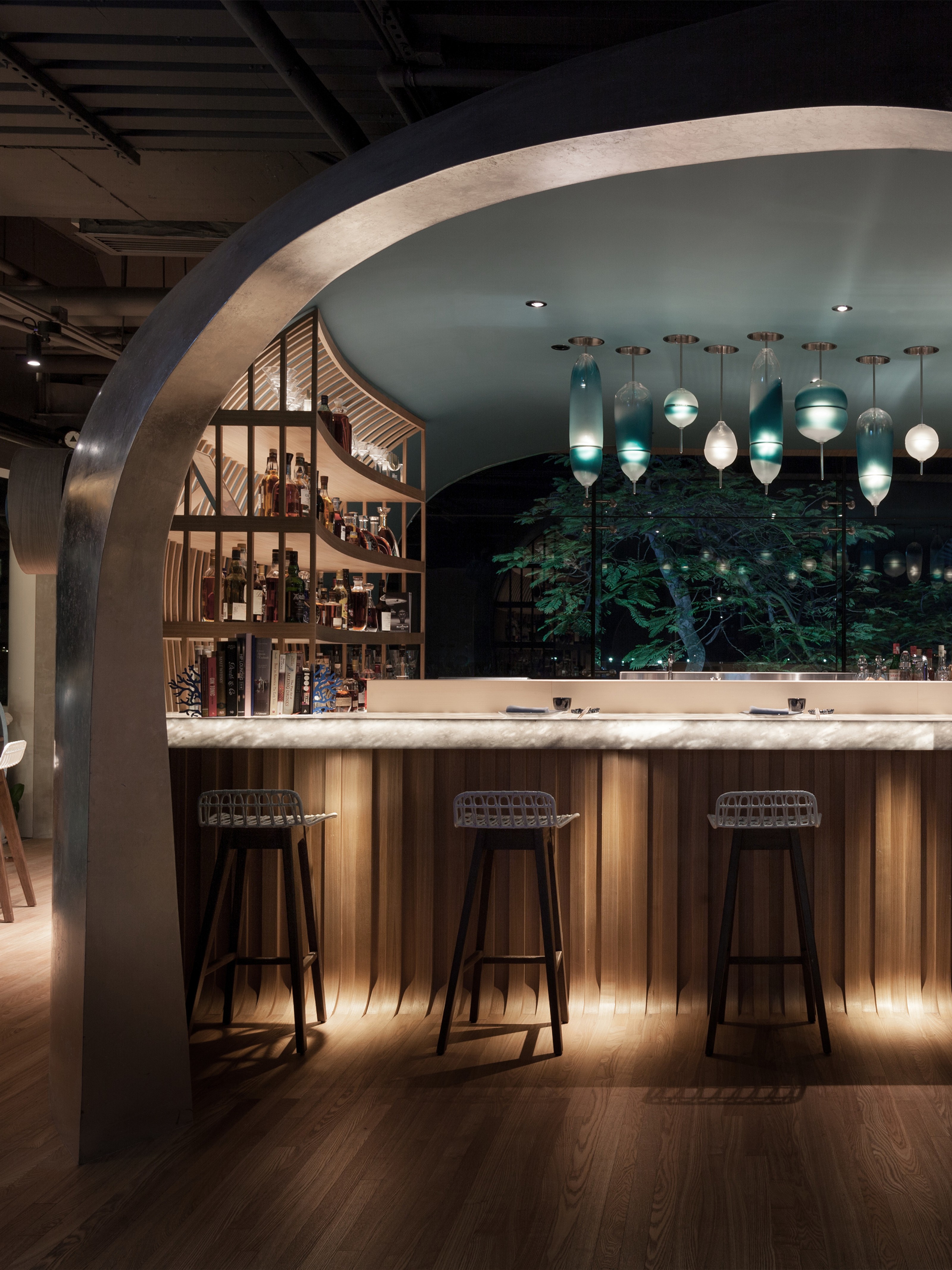 A stylish, modern bar with a curved archway, an illuminated marble counter, unique teal pendant lights, and a backlit liquor display, viewed at night.