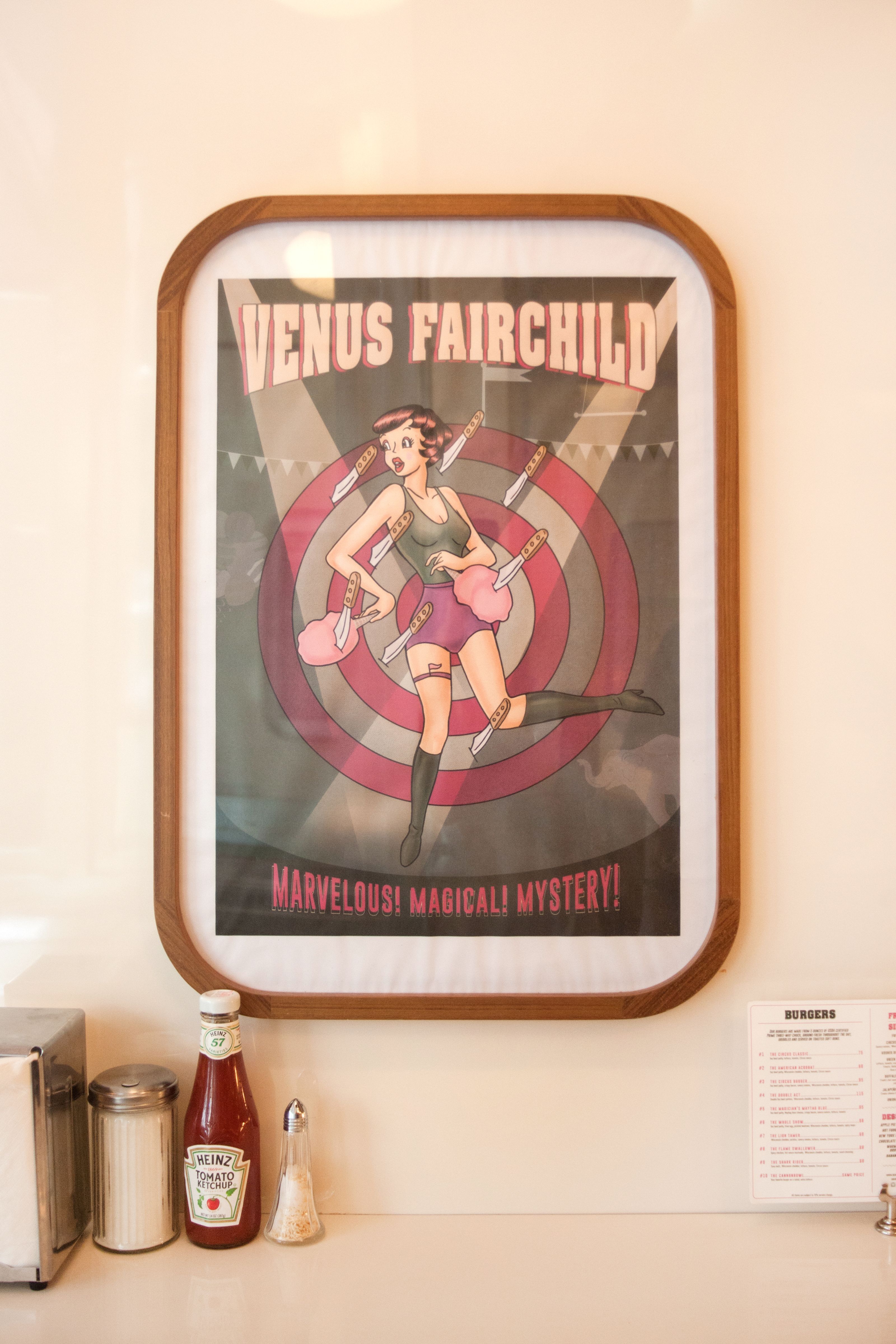 A framed retro circus poster titled "Venus Fairchild" shows a woman with knives stuck around her, above a diner counter with condiments.