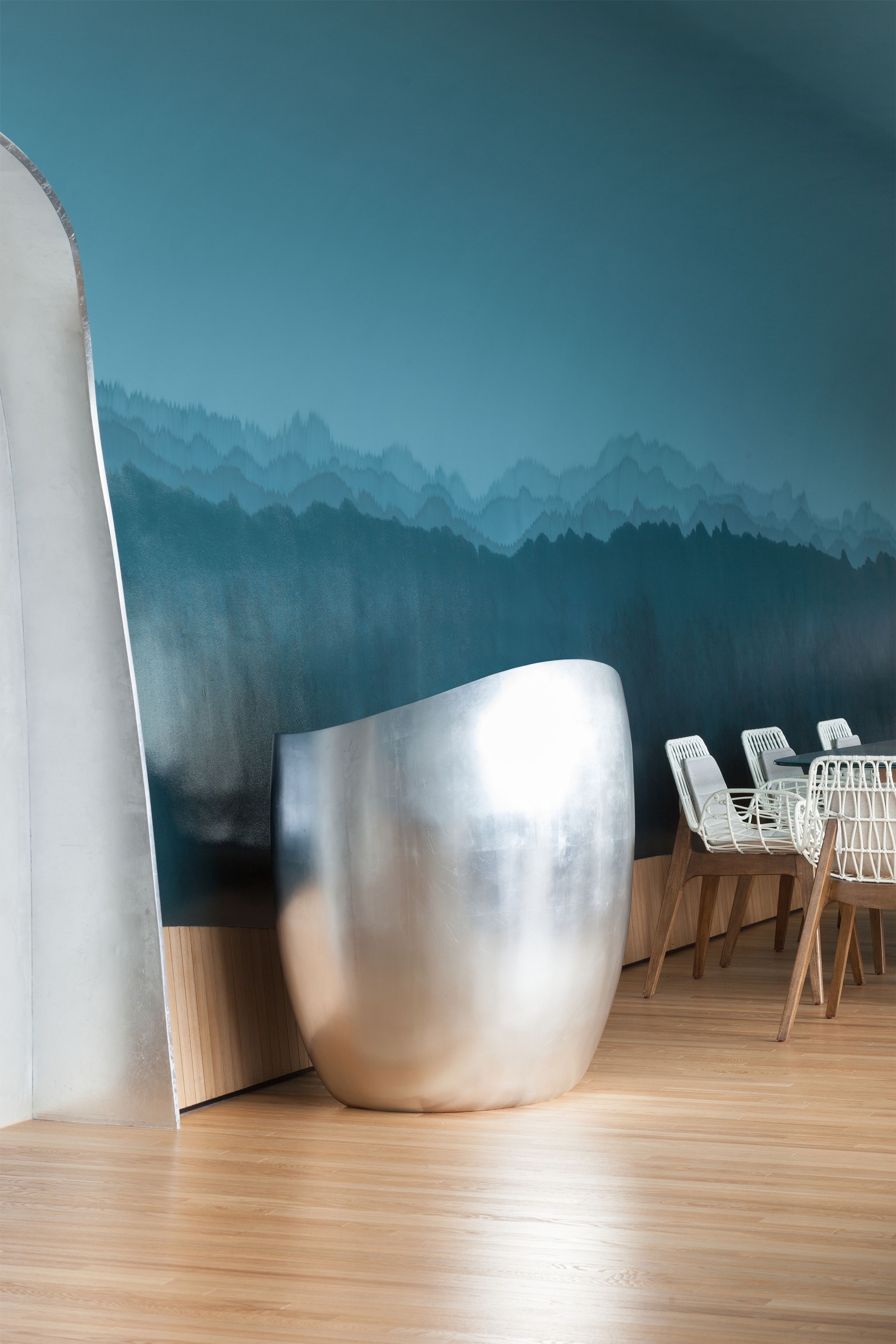 A modern room featuring a large, curved silver sculpture, a teal wall mural with layered mountains, white woven chairs, and a wooden floor.