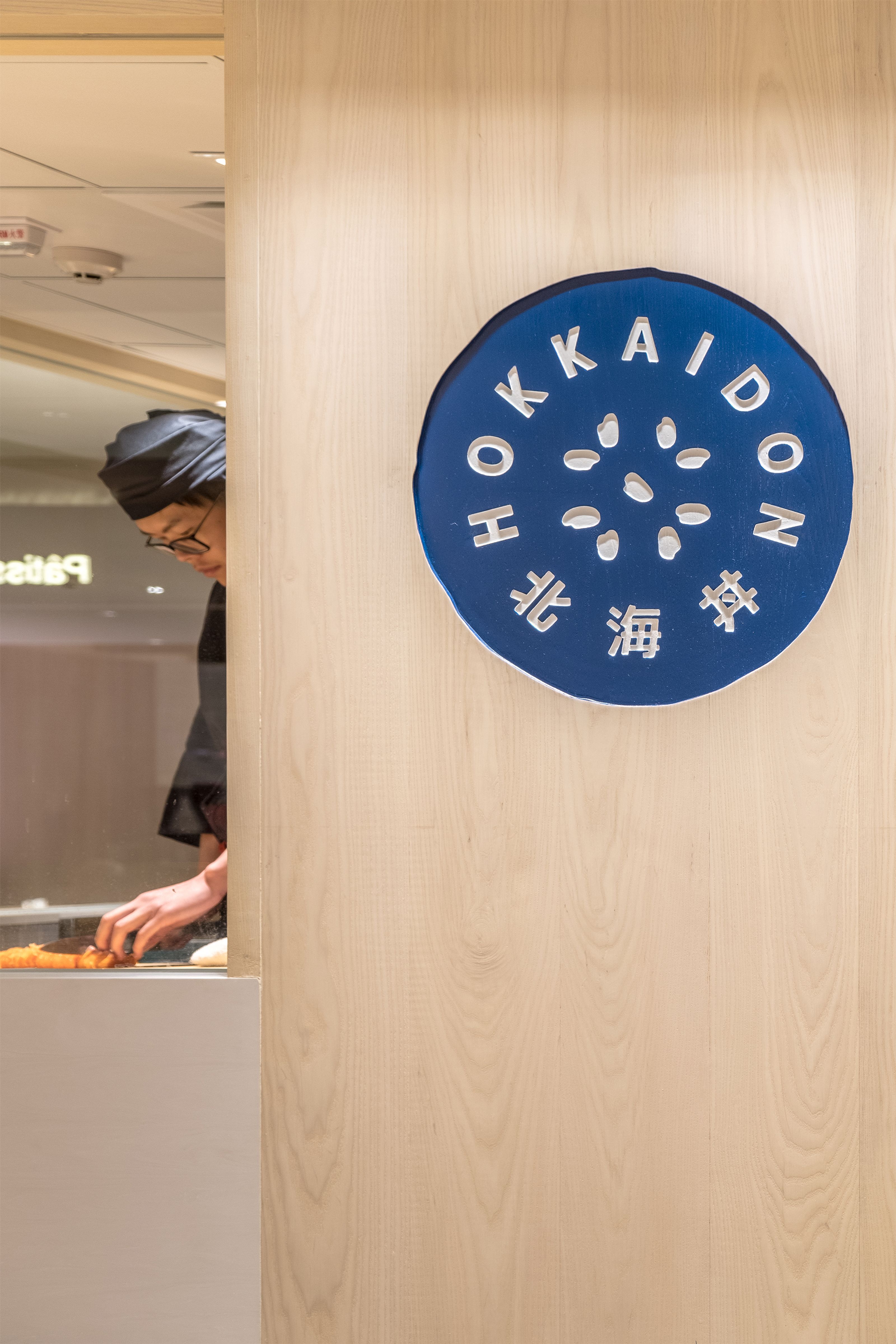 A chef in a black uniform prepares food behind a glass partition, next to a blue circular "HOKKAIDON" sign on a light wood wall.