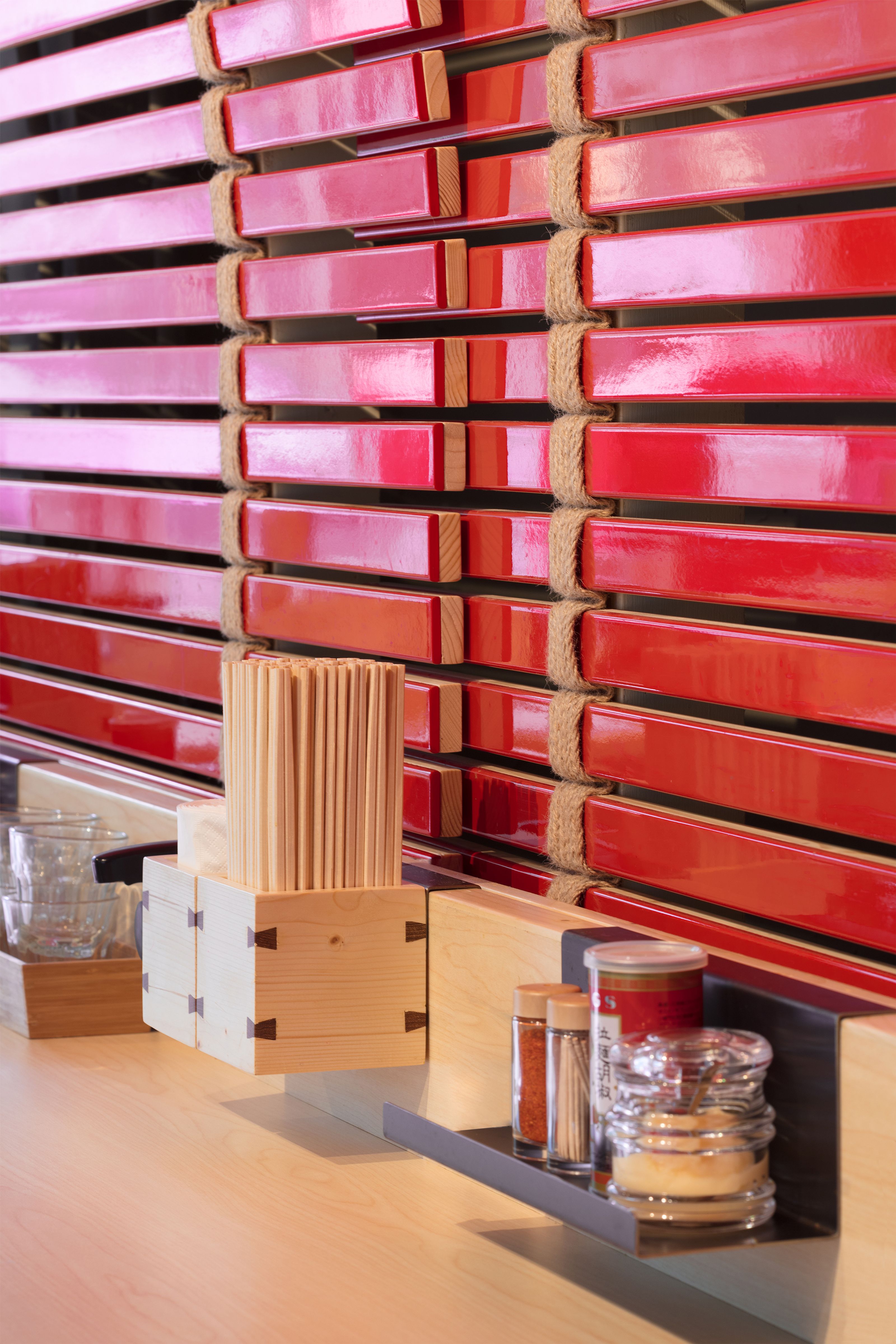 A wooden counter with a chopstick holder and spice shakers in front of a wall of red lacquered wooden slats.