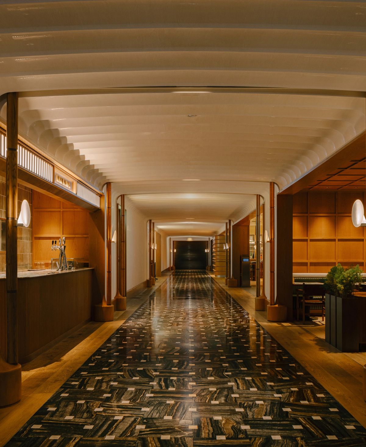 A long, luxurious corridor with a dark, patterned stone floor, warm wood-paneled walls, and a bar area on the left.