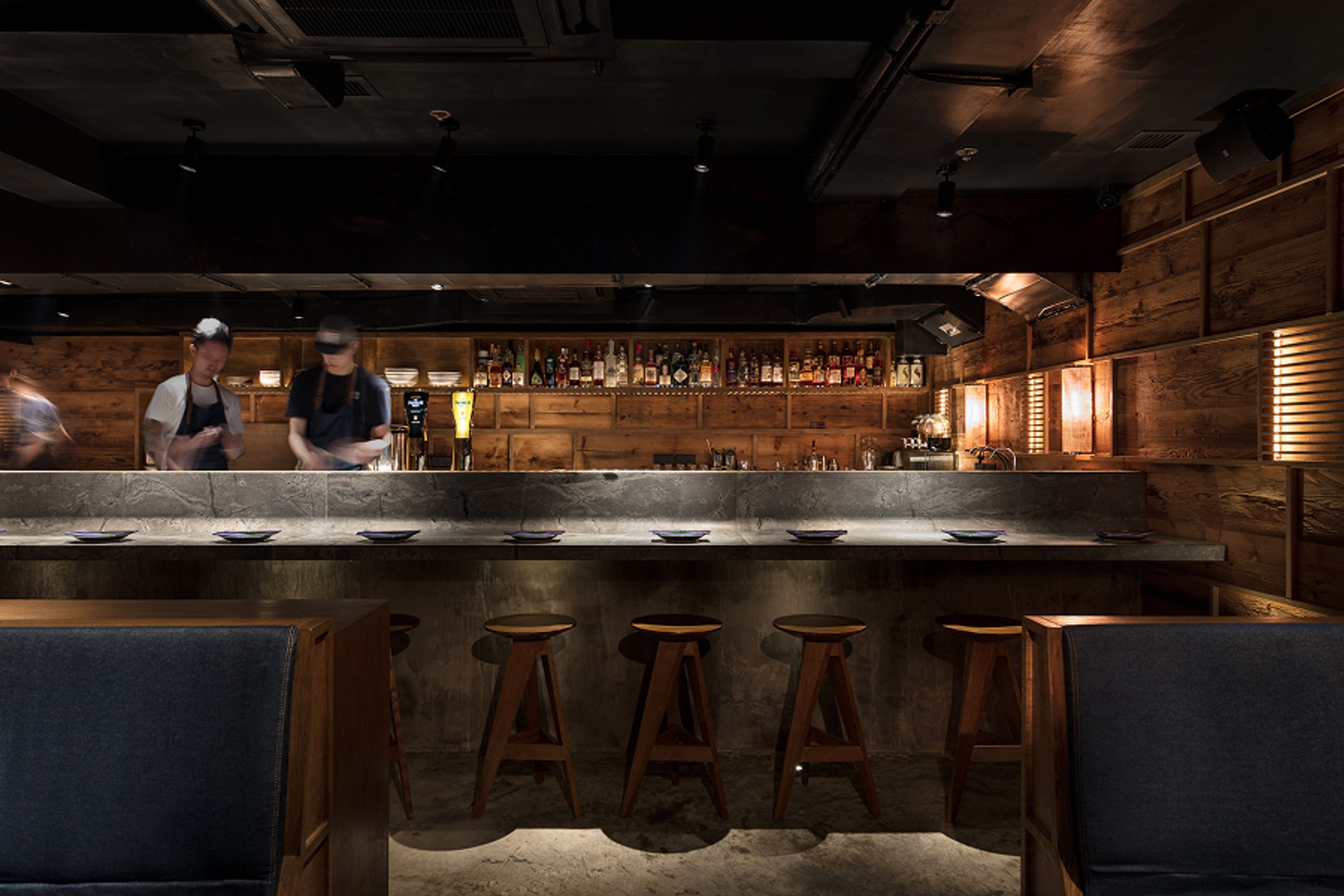 Modern, dimly lit restaurant bar with a concrete counter, two blurry staff, wooden crate walls, and bar stools.