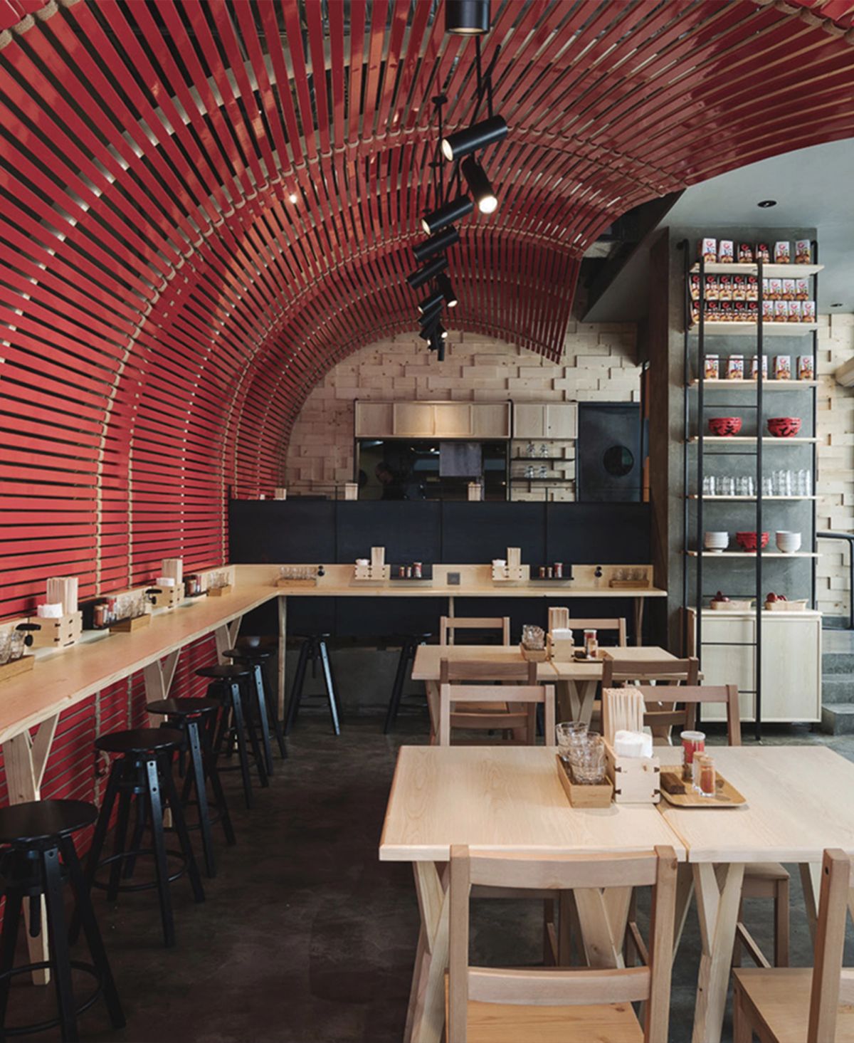 Modern restaurant interior with a vaulted red slatted ceiling, light wood tables, and black stools.